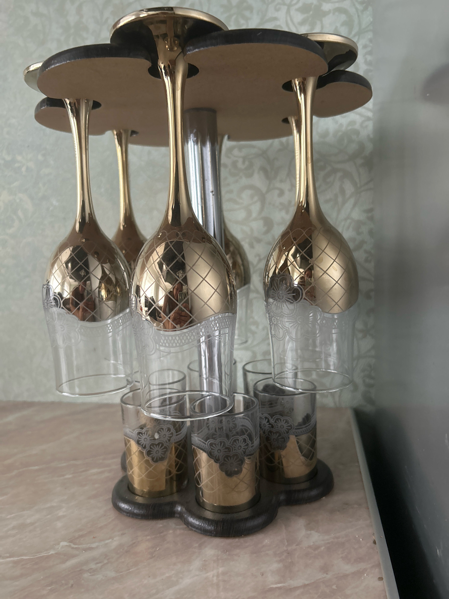 The image displays a gold and clear glass set on a marble surface. The set includes multiple stemmed glasses with gold-plated tops decorated with a diamond pattern and floral etching near the base, held upside down in a wooden rack. Below these, several smaller shot glasses with gold interiors and similar floral and diamond patterns are also housed in the rack. The background features a light green, patterned wallpaper and a metallic gray object on the right. The scene is indoors, likely in a domestic setting in Novopokrovskaya, Russia. No people are directly visible, though reflections in the glasses suggest their presence during the photograph's capture.