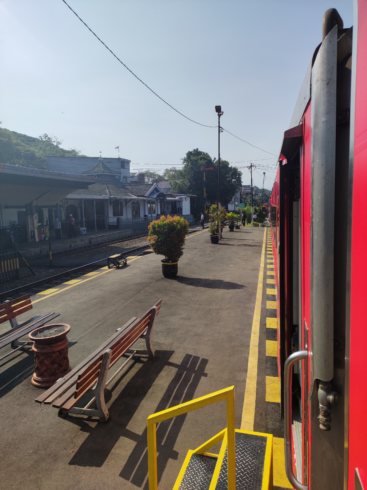 The image shows a train platform in Malang, Indonesia. A red train is stopped at the platform, with its doors open. The platform is paved with asphalt and has yellow and black checkered lines to mark the safety zone. There are several benches on the platform, some with wooden seats and metal frames. In the background, there are buildings with traditional tiled roofs, trees, and a clear blue sky. A few people are visible on the platform, some standing and some walking. The scene appears to be a train station, possibly during the day.