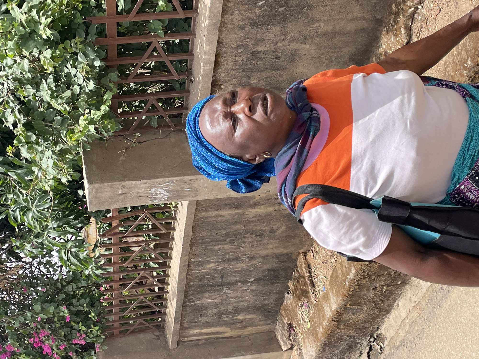 A middle-aged, dark-skinned woman wearing a blue headscarf, gold earrings, and a striped orange and white top is walking on a paved street in Jos, Nigeria. A black shoulder bag is slung over her shoulder. She is looking slightly upward with her mouth open. To her left is a concrete structure with a brown metal trellis covered in green foliage. The street surface is cracked and worn. The overall lighting suggests it is daytime.