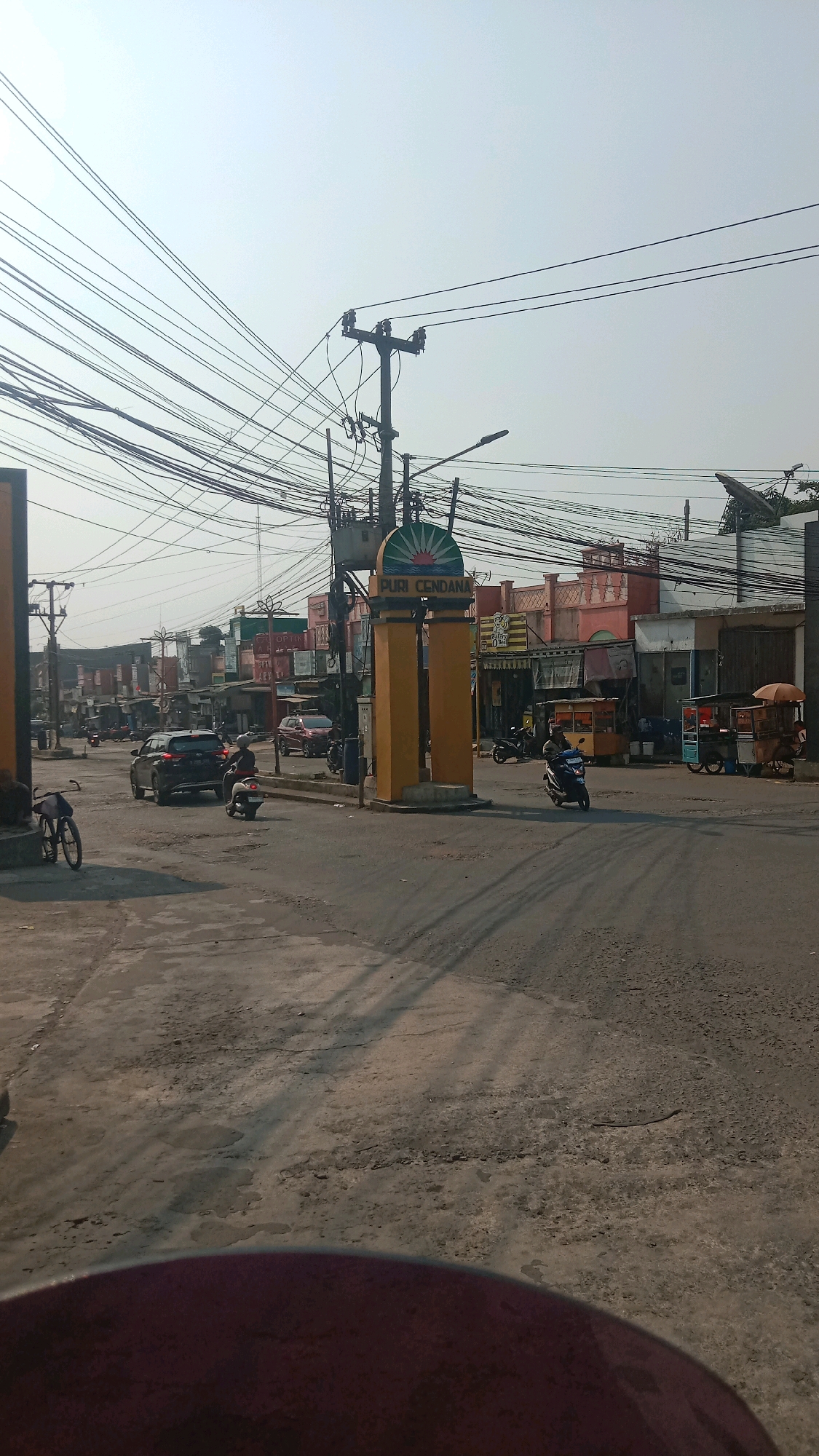 The image shows a street scene in Sumberjaya, Indonesia. A yellow archway with the words "PURI CENDANA" is prominently displayed. The street is paved with asphalt and has several vehicles, including cars and motorcycles. A bicycle is parked on the left side of the road. There are buildings lining the street, some of which appear to be shops. Overhead, a dense network of power lines crisscrosses the sky. The sky is bright and appears hazy, suggesting it is daytime. There are a few people visible on motorcycles and in the shops. The overall impression is of a busy commercial street in a town or city.