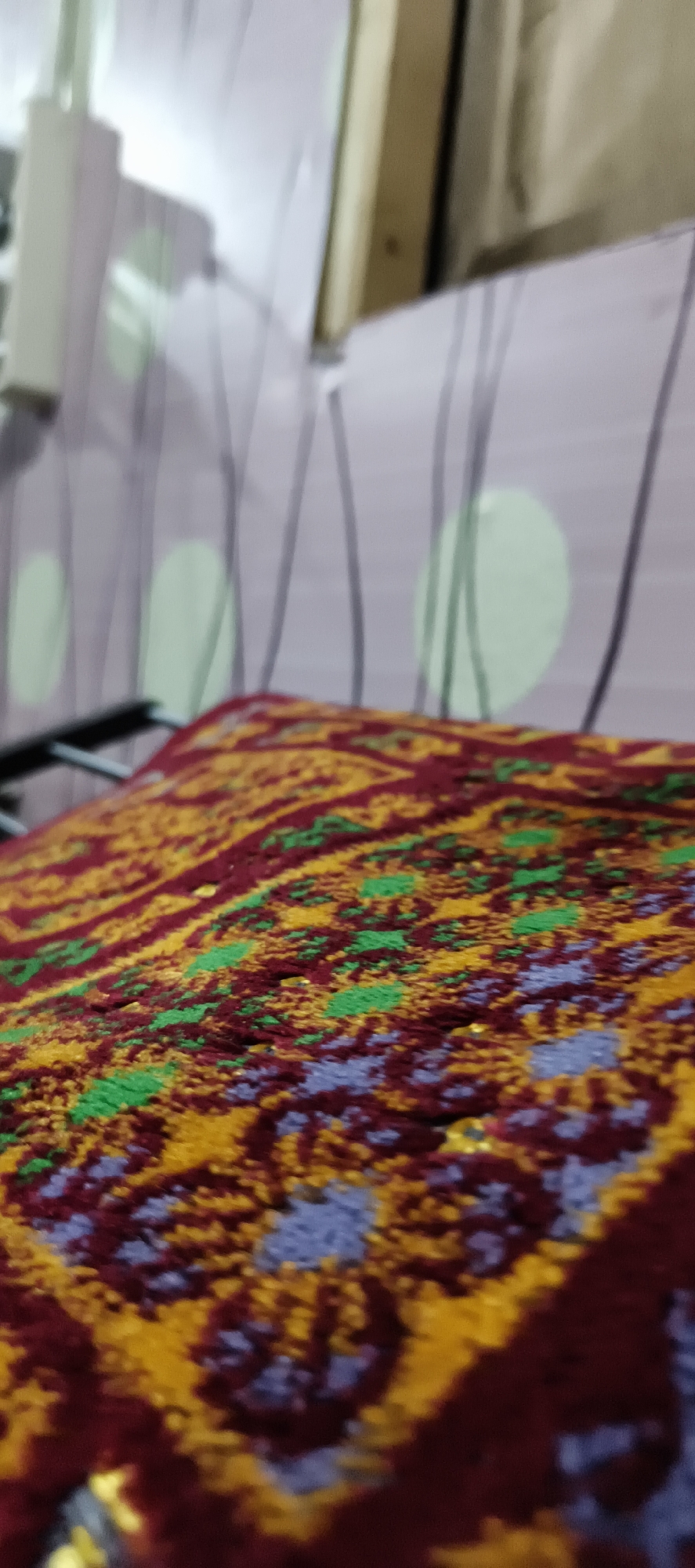 The image shows a close-up view of a patterned textile, likely a rug or blanket. The pattern features intricate geometric designs in shades of burgundy, yellow, green, and a muted blue or purple. The textile is draped over a dark, possibly metal, frame. In the blurred background, a wall with a subtle striped pattern and oval shapes in a light purple and green hue is visible. A portion of a wooden structure and a white rectangular object, possibly a light fixture or part of a wall, are also present. The scene appears to be indoors, in a residential setting. There are no people or animals visible in the image. The location is identified as Muaro Jambi, Indonesia.