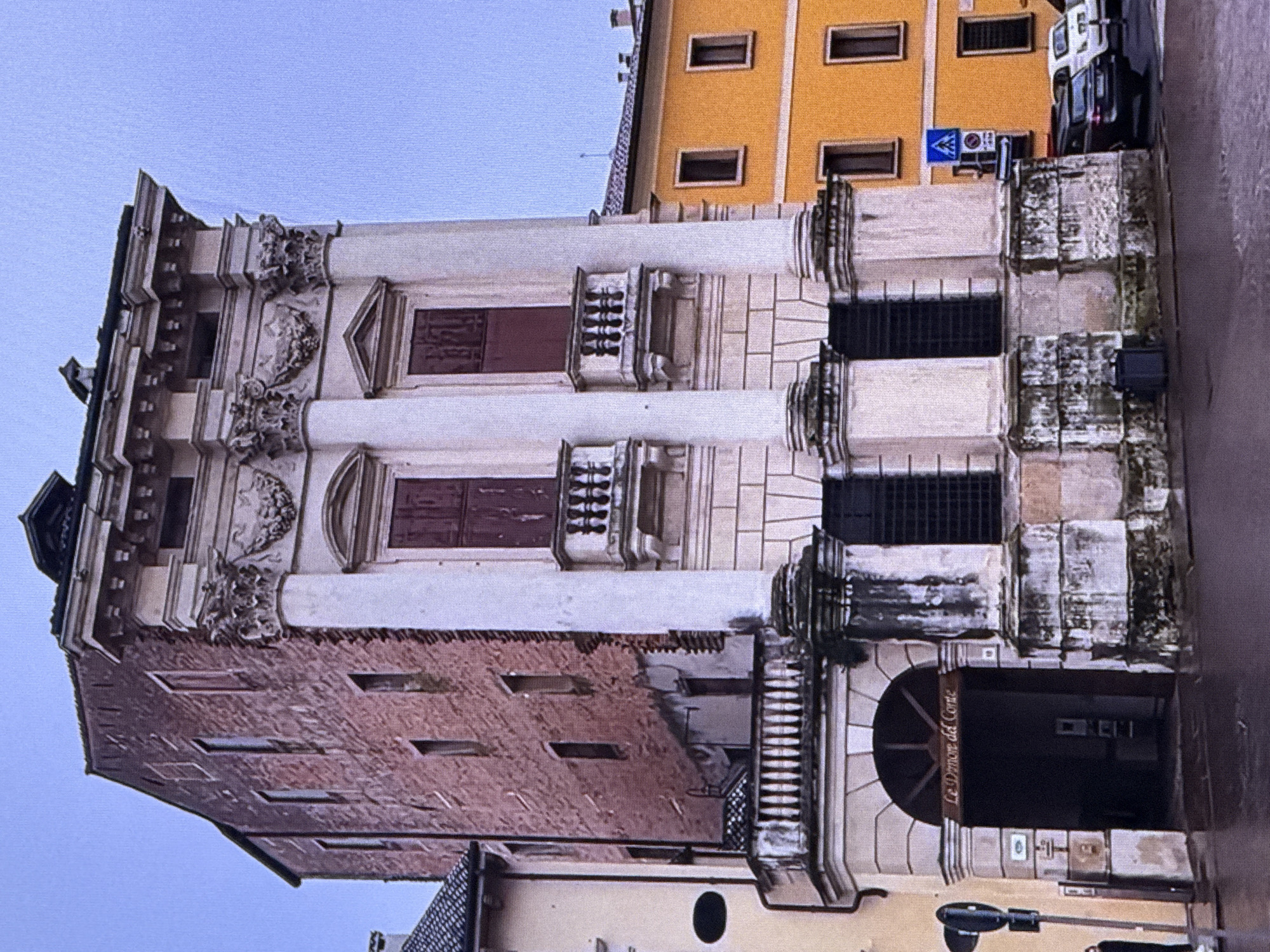 The image depicts a historical building with a brick facade on the left and a more ornate, lighter-colored section with classical architectural elements on the right. The ornate section features large columns, balconies with decorative railings, and arched doorways. Adjacent to the main building on the right is a bright yellow building with rectangular windows. The street in front of the buildings appears wet, suggesting recent rain. Several cars are parked along the curb in front of the yellow building. There is a traffic sign with a blue triangle and a white 'A' on a pole near the cars. A person is partially visible on the far left, standing near a dark object. The sky is overcast and a pale blue. The overall impression is of an old European city with a mix of architectural styles. This location is not in Dubai, United Arab Emirates.