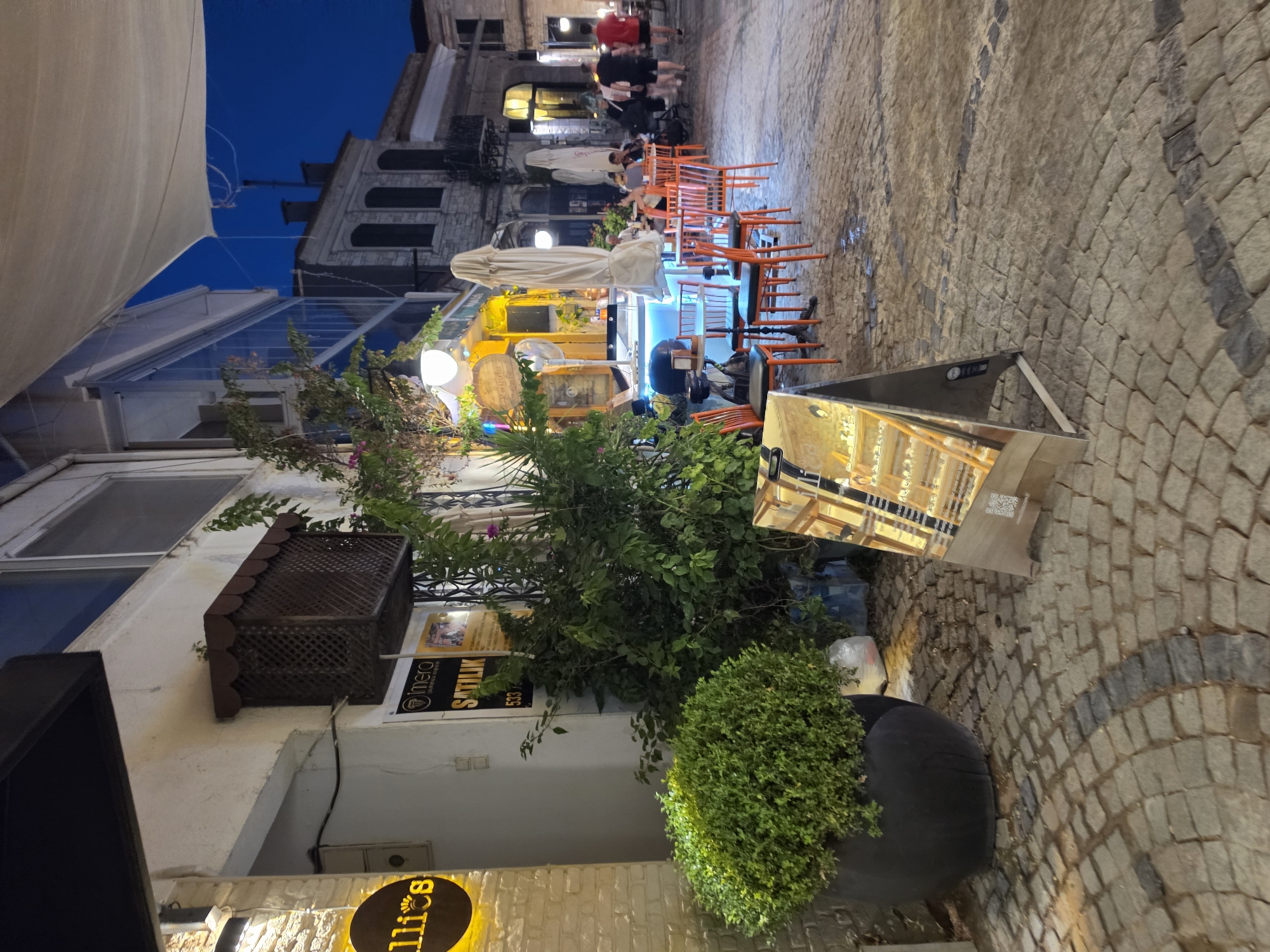 The image depicts an evening scene on a cobblestone street in Urla, Turkey. A cafe or restaurant with outdoor seating is visible, featuring orange chairs and tables. A large white umbrella shades part of the seating area. A decorative plant in a large black pot is positioned in front of the building. Above the plant, a brown wooden lattice structure is mounted on the wall.
In the background, several people are seen walking on the street. The buildings lining the street have traditional architecture. An awning, possibly for shade or rain protection, extends from one of the buildings. The sky is dark blue, indicating it is either dusk or early evening. Artificial lighting from the cafe and surrounding buildings illuminates the scene.
On the wall of the building, there are signs. One sign is yellow with black text that appears to read "SATILIK" (which means "for sale" in Turkish) and the number "533" below it. Another circular sign with yellow text on a black background is partially visible at the bottom left. A reflective triangular object, possibly a display stand or part of a store, is present on the right side of the image, showing reflections of the street and buildings. The overall atmosphere suggests a lively but relaxed evening on a charming street.