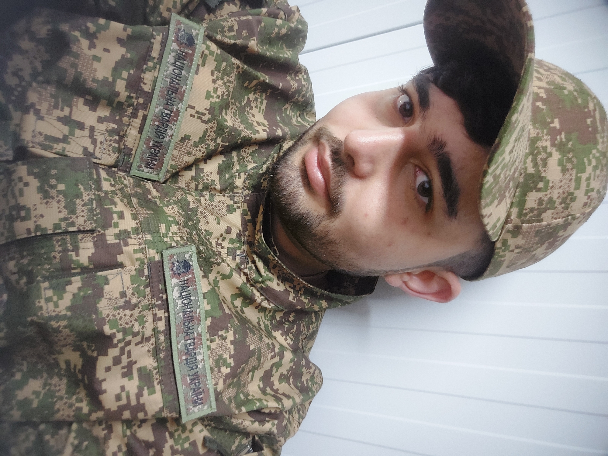 A close-up photograph shows a man wearing a camouflage uniform and matching cap. The uniform has a patch on the chest and sleeve with text in Ukrainian, which translates to "National Guard of Ukraine". The background is a white wall with horizontal panels. The image does not contain enough information to determine the location in the city of Huliaihorodok, Ukraine.