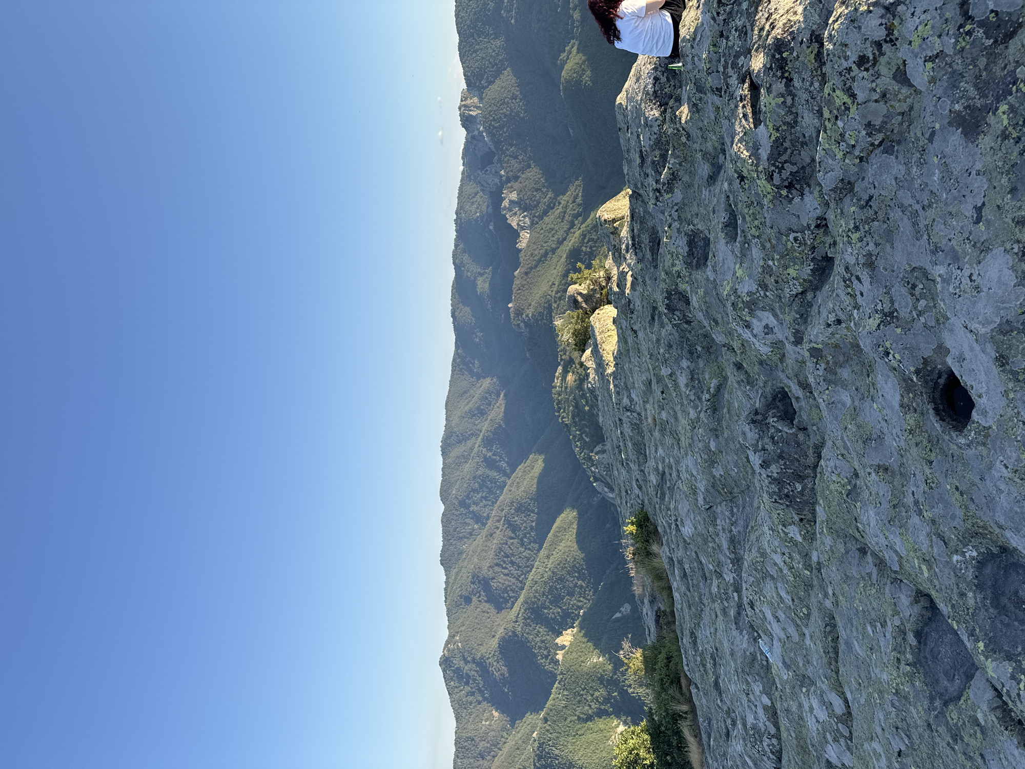 The image shows a scenic view of the Vitosha mountain range near Sofia, Bulgaria, specifically capturing the landscape of Sini Vrah. The foreground is dominated by rugged, weathered grey rocks covered in patches of green lichen. In the mid-ground, a person with dark hair, wearing a white t-shirt and dark bottoms, is seated on the rocks, facing away from the camera and looking towards the mountains. Beside them, a green bottle is visible. Beyond the rocky terrain, the image opens up to a vista of rolling, forested mountains stretching into the distance under a clear, pale blue sky. The sunlight casts shadows across the mountain slopes, highlighting the undulating terrain.