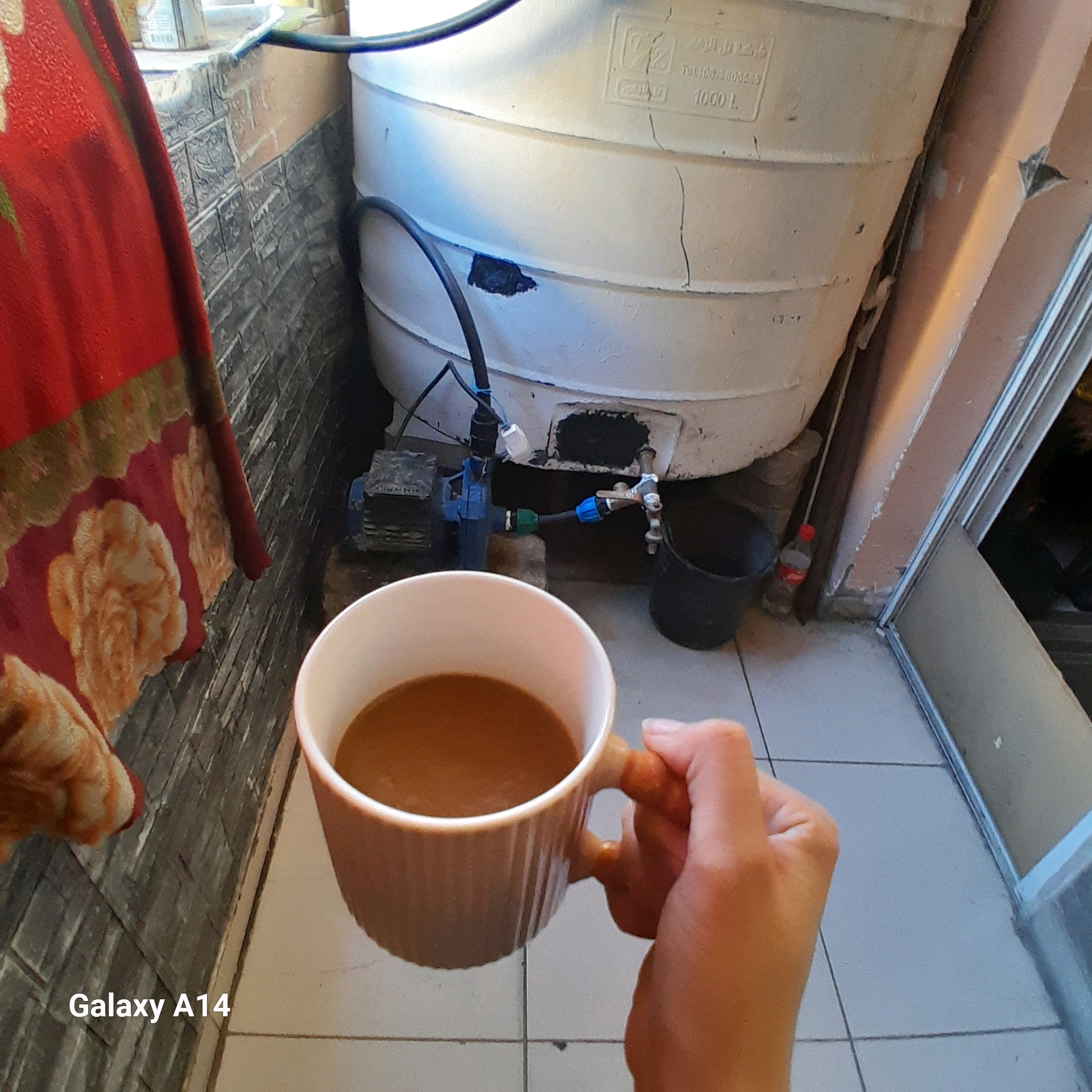 A person's hand holds a tan mug filled with a brown liquid, presumably coffee, in the foreground. The mug has a ribbed texture and an orange-brown handle. Behind the mug, a large, white, ribbed plastic water tank with a capacity of 1000 liters is visible. The tank has some visible cracks and a dark patch that appears to be a repair. Attached to the tank is a blue water pump and tubing. A black bucket is situated near the base of the tank. To the left of the tank and pump, a red fabric with a floral pattern hangs down. The background consists of a tiled wall and floor. The overall scene appears to be an outdoor or semi-outdoor area, possibly a yard or balcony, in Khan Yunis, Palestinian Territories. A watermark at the bottom left reads "Galaxy A14".