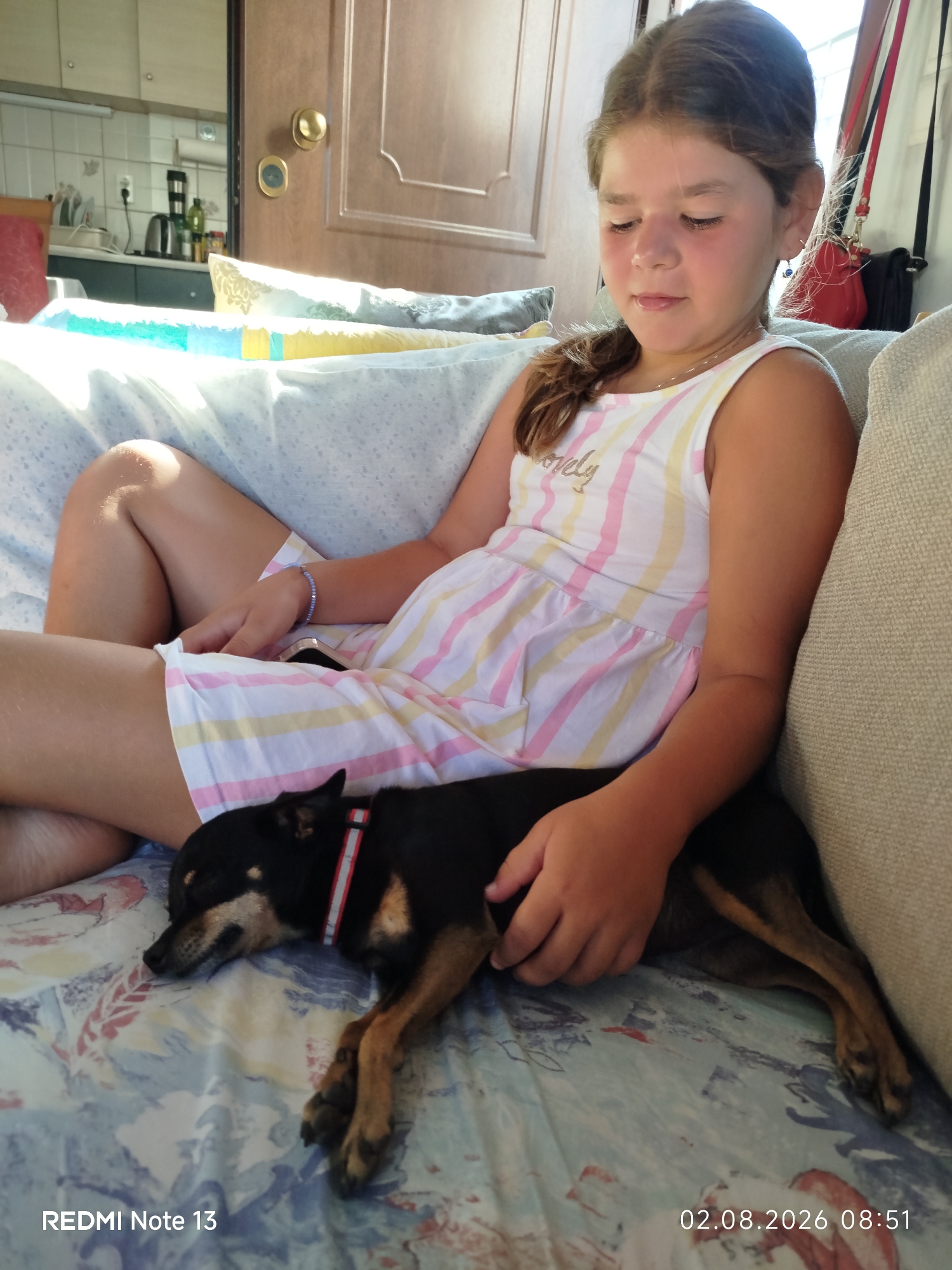 A young girl is sitting on a couch with a small black dog sleeping on her lap. The girl is wearing a striped dress and has a bracelet on her wrist. The dog is wearing a red and white collar. The couch is covered with a patterned blanket. In the background, a kitchen is visible through an open doorway. The scene appears to be indoors, possibly in a home. The image includes a timestamp in the lower right corner indicating the date as August 2, 2026, and the time as 08:51.