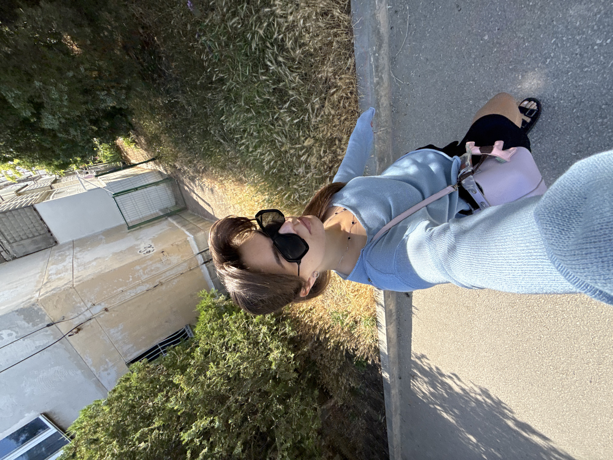 A young woman with brown hair, wearing sunglasses, a light blue sweater, black shorts, and sandals, walks down a sidewalk. She carries a pink purse. To her left is a building and trees. To her right is overgrown grass. The sky is visible in the background.