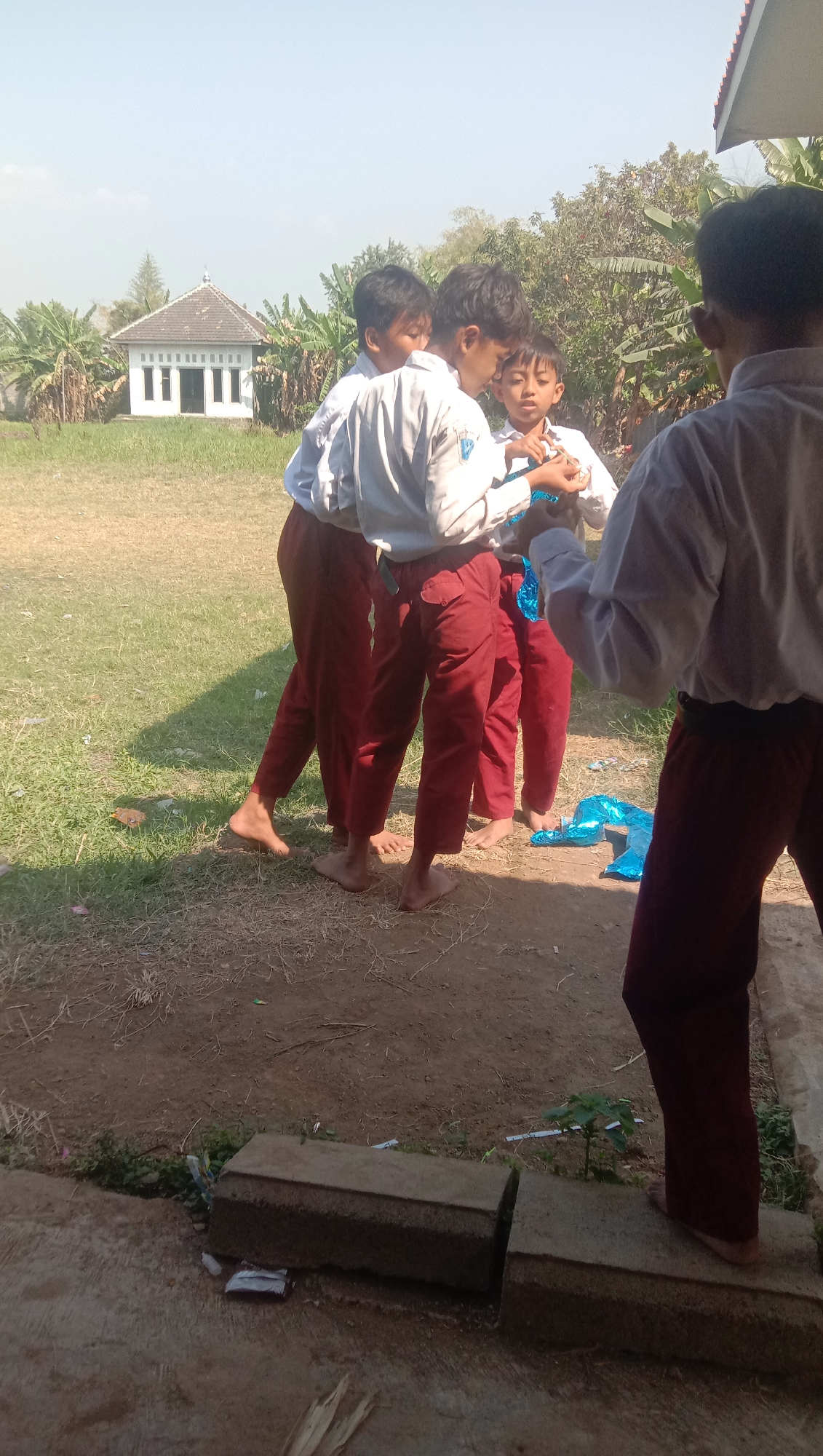 Four children, likely elementary school age and wearing school uniforms consisting of white shirts and maroon pants, are gathered outdoors. Three children are in the foreground, appearing to interact with each other, possibly playing with a blue balloon object on the ground. The fourth child is partially visible on the right, also in uniform. The environment is grassy with patches of dry earth and vegetation. In the background, there's a small white building with a distinctive roof, resembling a mosque or community hall, surrounded by trees and banana plants. The sky is clear and bright, suggesting a sunny day. The ground in the immediate foreground shows a concrete structure with some debris. The scene is likely a schoolyard or public area in Karang Anyar, Indonesia, given the architectural style of the building and the presence of banana plants, which are common in tropical regions.