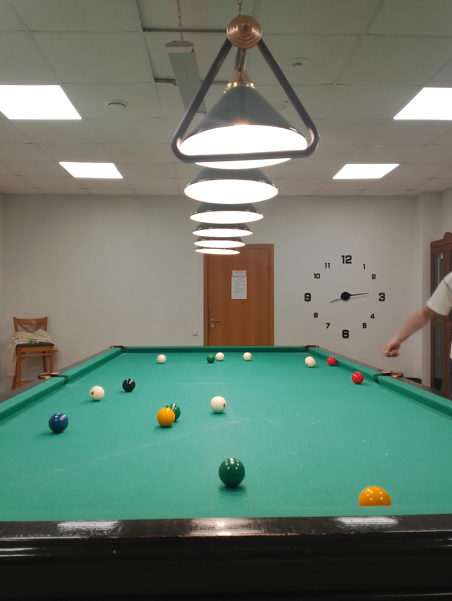 The media file depicts a billiard room, likely in Izhevsk, Russia. The scene is dominated by a large, green billiard table with several balls scattered across its surface. The balls are of various colors, including white, black, blue, red, green, and yellow, some of which appear to be in play. The room is well-lit by fluorescent ceiling lights and a prominent, multi-tiered chandelier hanging over the billiard table.
On the wall behind the table, a large, modern clock with black numerals and hands indicates the time is approximately 2:45. A wooden door is visible to the right of the clock, and further to the right, a portion of a wooden cabinet is present. To the left of the billiard table, a wooden chair with a beige cloth draped over it is placed against a pale blue wall.
A person's arm and shoulder are visible in the lower right corner of the frame, suggesting they are either playing billiards or observing the game. Their skin tone and the casual attire (white t-shirt) do not provide strong clues about the time of day or weather. The overall atmosphere of the room appears casual and recreational. There is a small informational poster visible on the door.