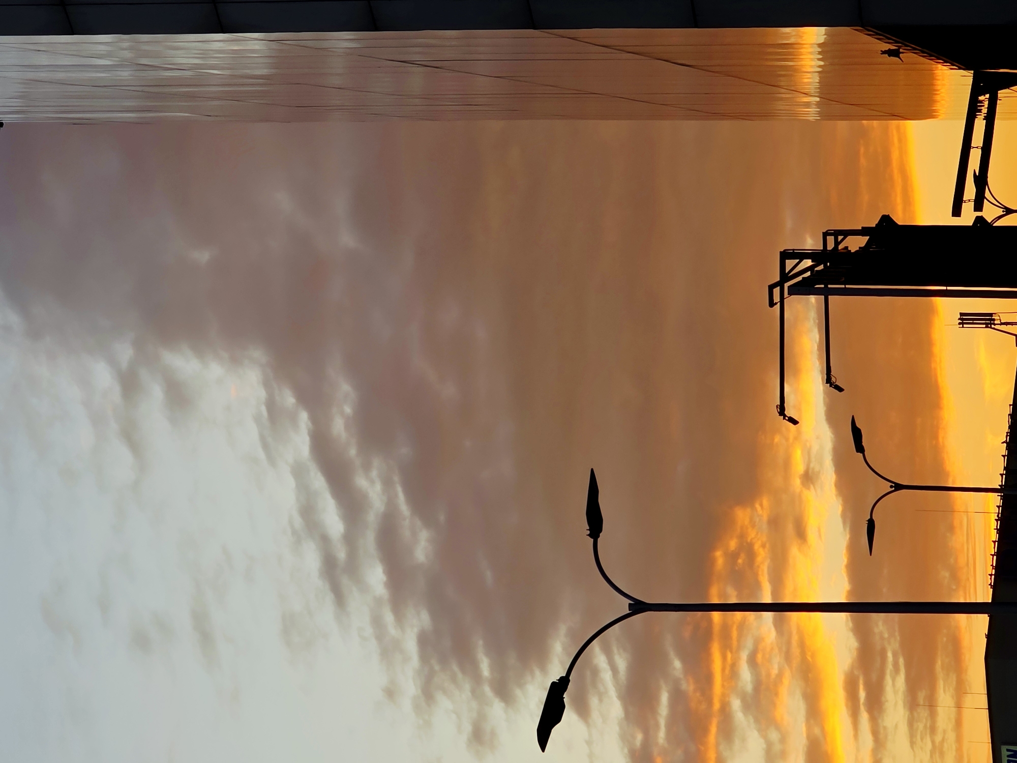 A street lamp is silhouetted against a vibrant orange and yellow sunset sky. The sky is filled with dramatic clouds that are illuminated by the setting sun. To the right of the street lamp, a tall, modern building with a reflective glass facade rises into the sky. The building's facade mirrors the colors of the sunset, creating a stunning visual effect. The overall scene captures the beauty of a sunset in an urban environment, with the artificial lights of the city starting to compete with the natural light of the sky.