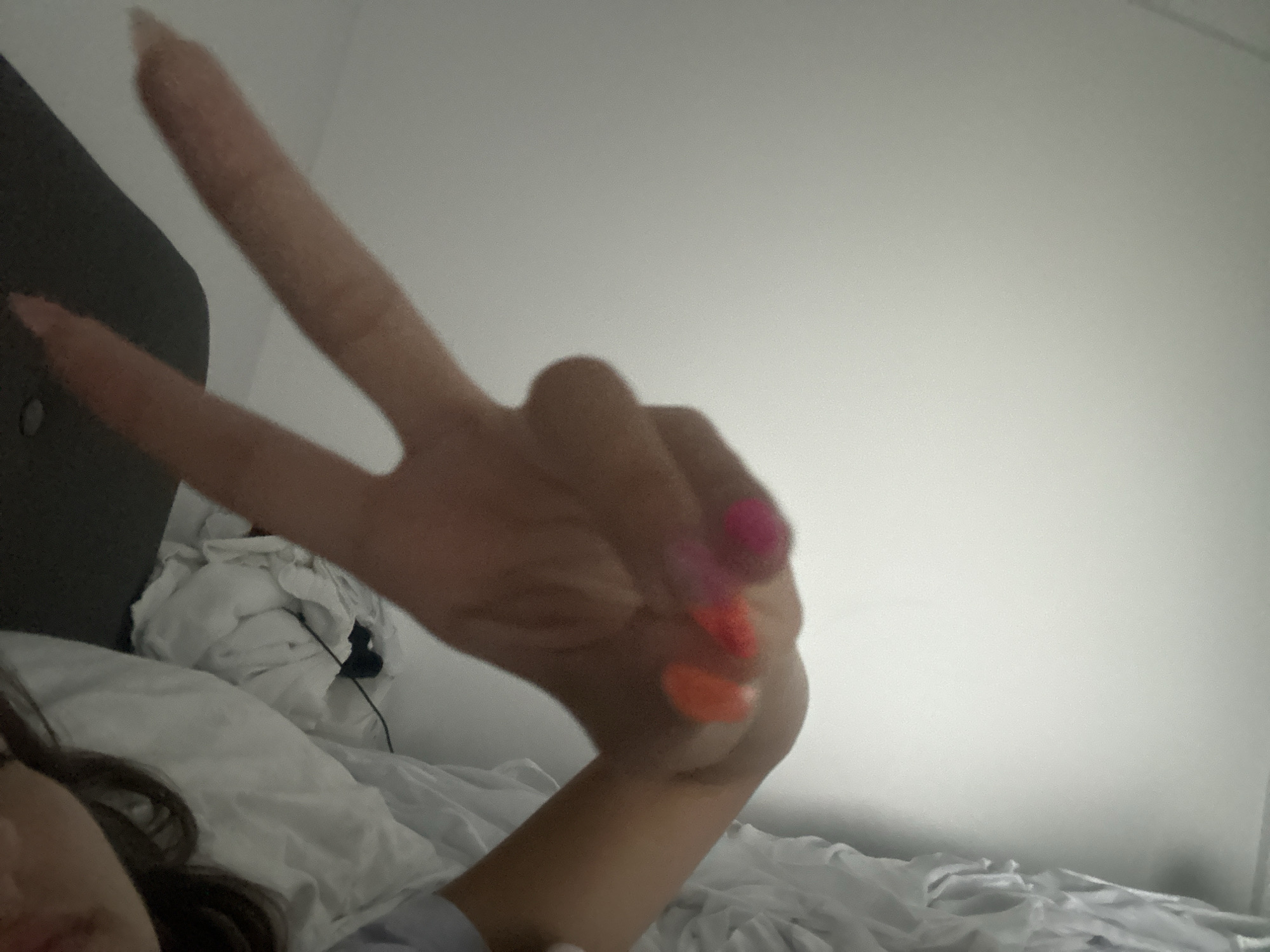 The image depicts a close-up view of a person's hand forming a peace sign, with the index and middle fingers extended upwards. The hand has brightly colored nail polish, with shades of pink, orange, and red visible. The background consists of white bedding and a blurred-out wall. The person appears to be lying down, possibly in a bed, with the hand positioned in the foreground. The scene is set in Stațiunea Zoologică Marină Agigea, Romania.