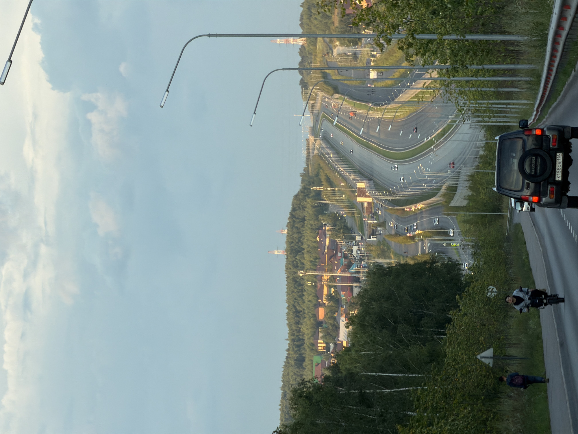 The image depicts a winding highway with several lanes of traffic, curving through a suburban landscape. In the foreground, a black Suzuki Jimny SUV is driving on the right lane of the highway. A person is riding a motorcycle on the shoulder of the road to the left of the SUV. Further down the highway, other vehicles are visible, including cars and a white minivan.
On the left side of the highway, a dense green forest lines the road, interspersed with trees and shrubs. Beyond the forest, a suburban area with houses featuring red and orange roofs is visible on a gently sloping hill. In the distance, two tall red and white radio towers stand out against the sky. The sky is a pale blue with scattered white clouds. The scene suggests a bright, possibly late afternoon or early evening, setting. The location is identified as Kazan, Russia.