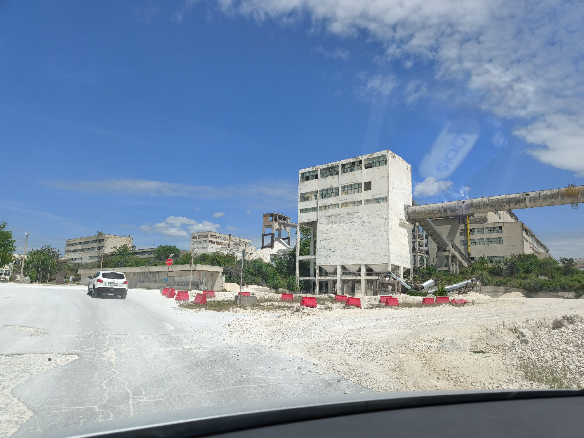 The image depicts an industrial landscape with a large, white, multi-story building possibly located near a quarry or processing plant. The building is rectangular with multiple windows and appears to be connected to a large conveyor belt system. The ground around the building is covered in light-colored material, possibly crushed stone or limestone. Red construction barriers are visible in front of the building. In the background, another building can be seen. A white SUV is driving on a road towards the viewer. The sky is clear blue with scattered clouds. The view is taken from inside a vehicle. The location and city are Unknown.