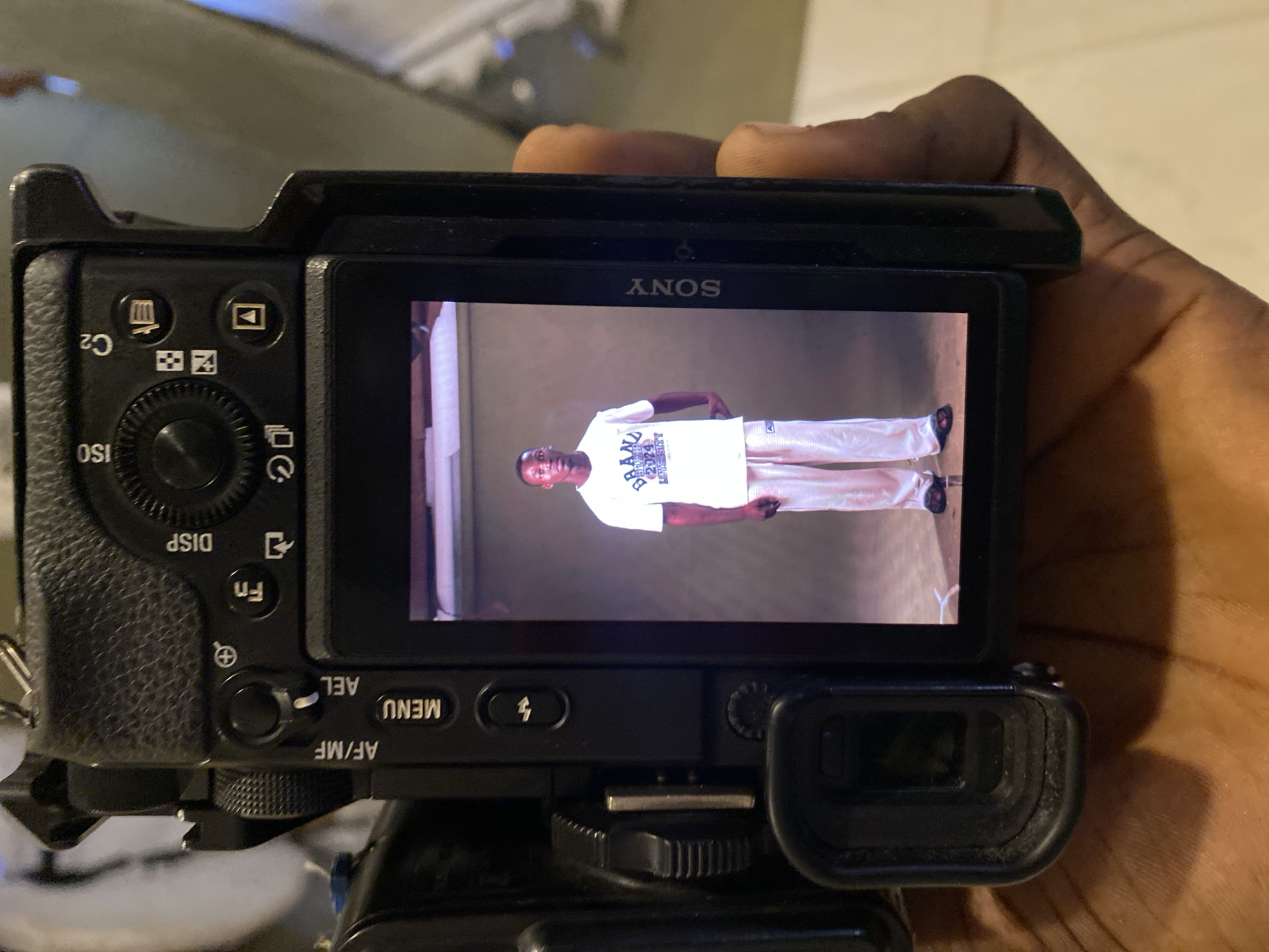A close-up shot captures a black Sony mirrorless camera being held by a person's hand, viewing an image on its rear LCD screen. The camera is positioned horizontally, while the image displayed on the screen is a vertically oriented full-body portrait of a young man.
The man in the photo stands facing forward, wearing a white t-shirt and light-colored (possibly white or off-white) pants with black shoes. Text is visible on his t-shirt, which appears to read "BRAN UNIVERSITY 2024 LOVE SITY" (some parts are indistinct). The background in the displayed image is a simple, light-colored wall, suggesting an indoor setting.
On the camera body, several controls and text labels are visible, including "SONY" at the top of the screen. Around the camera's rear, legible text includes "ISO", "C2", "DISP", "Fn", "AF/MF", "MENU", and "AEL", along with various function icons (playback, trash, flash, etc.). The camera appears to be on a tripod mount or similar support, partially visible at the bottom. The lighting in the scene is somewhat dim, suggesting an indoor environment, possibly in the evening or at night. The location context for this media file is Badagry, Nigeria.