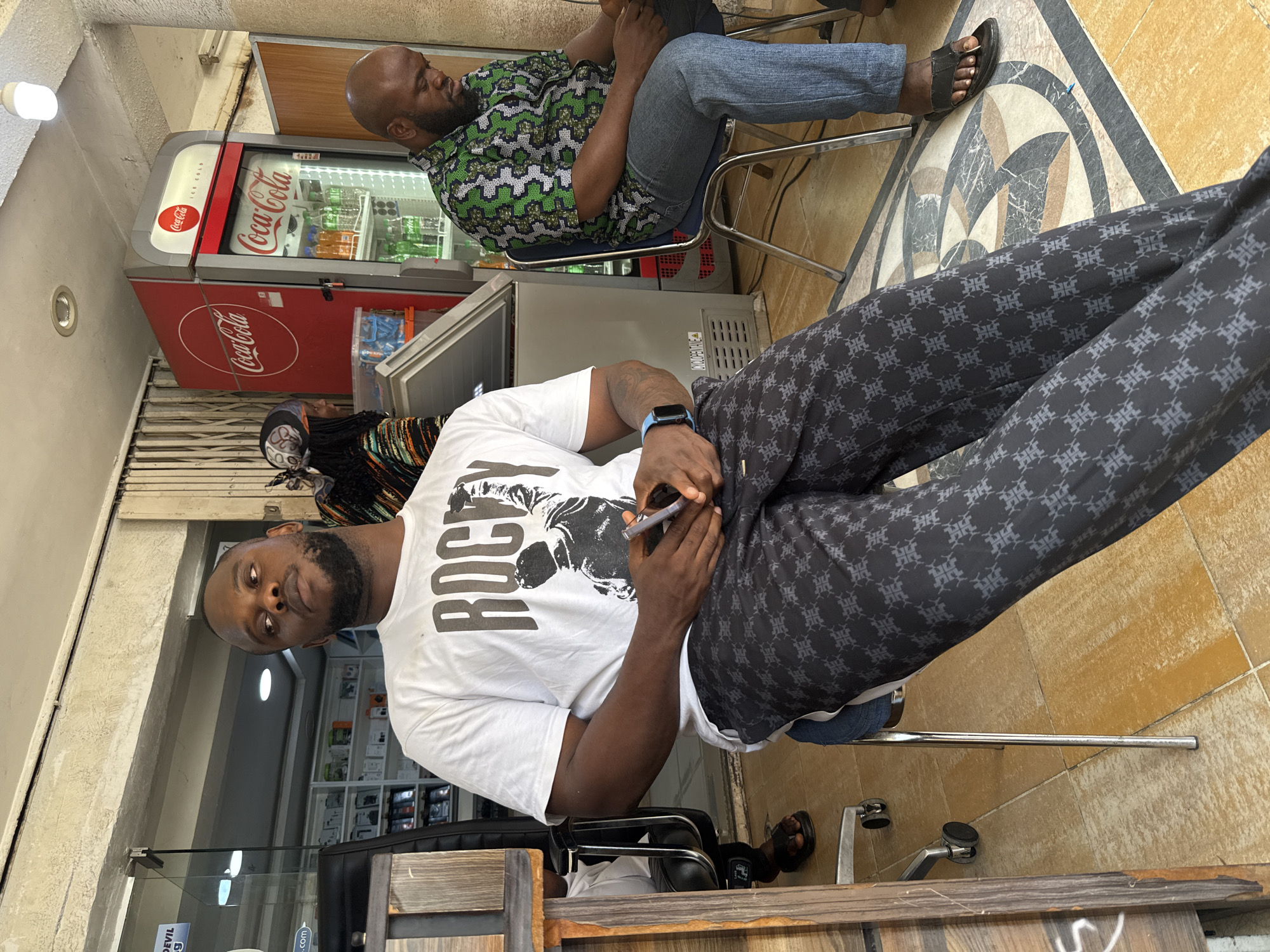 Two men are seated indoors, likely in a shop or waiting area in Abuja, Nigeria. The image is oriented sideways, but if viewed upright, one man is in the foreground, facing the camera, while the other is in the background, facing away.
The man in the foreground has dark skin, a short beard, and is wearing a white t-shirt with "ROCKY" printed on it, alongside a graphic of a figure. He wears dark pants with a light-grey repeating pattern and a blue smartwatch on his left wrist. He is holding a smartphone and a stylus/pen in his hands and is looking directly at the camera with a neutral expression. He is seated on a black chair with a metal frame, positioned behind a wooden counter or partition.
In the background, a second man with dark skin and a short beard is seated on a metal-framed chair. He wears a green, black, and white patterned shirt, blue jeans, and dark sandals. He is looking downwards, possibly at his own phone.
The setting includes a prominent red Coca-Cola refrigerator displaying "Coca-Cola" branding and "ICE COLD" text, stocked with various bottled beverages. Behind the man in the background, shelves filled with items are visible. The floor features a central section of patterned grey and white tiles, bordered by plain light brown tiles. Artificial lighting illuminates the space. Partially visible text on a sign near the bottom left (if upright) includes the word "DEVIL."