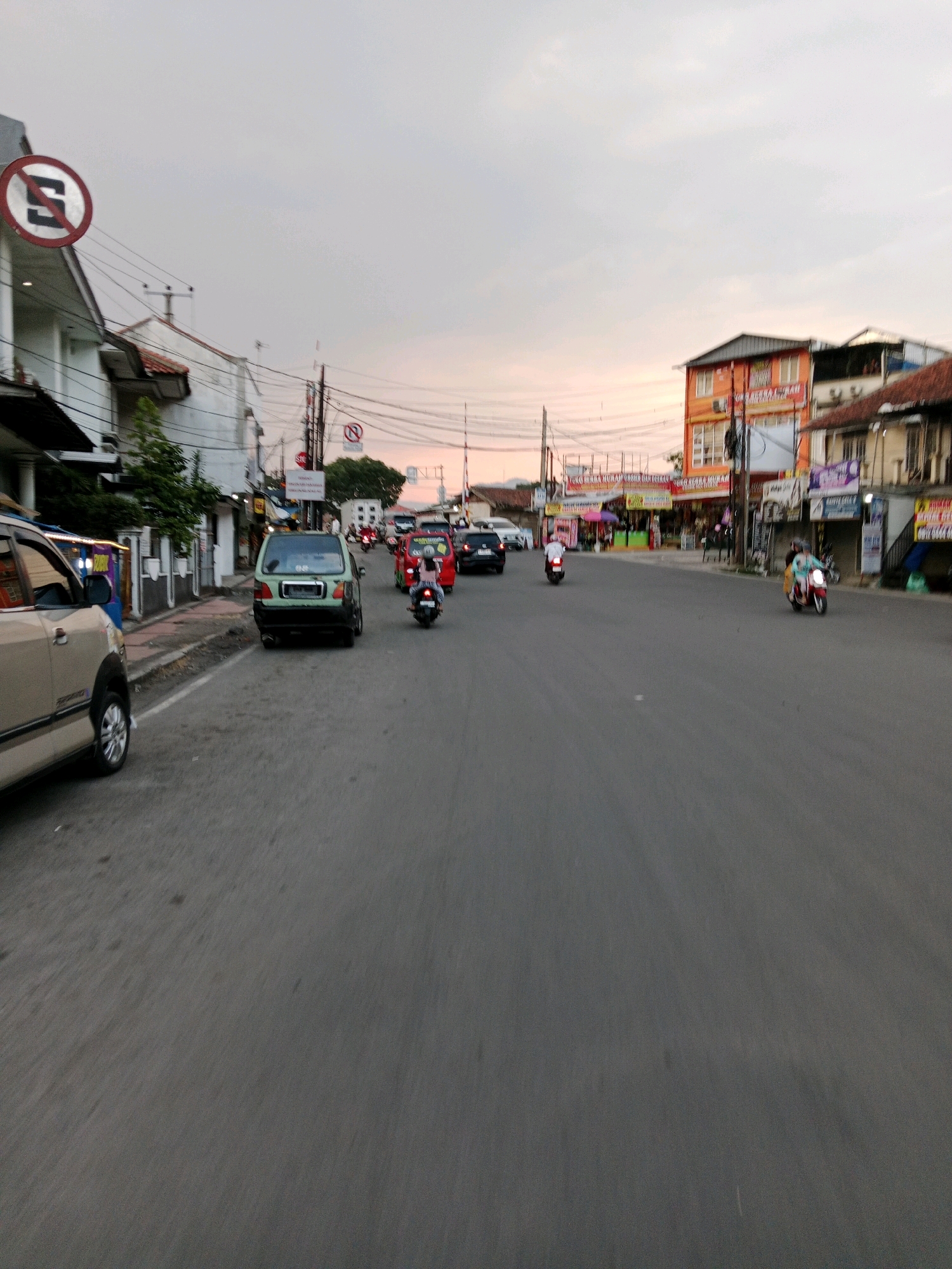 The image depicts a street scene in Sirnagalih, Indonesia, during what appears to be late afternoon or early evening, as indicated by the soft, diffused light and the colors in the sky. The road is paved and shows signs of traffic, with several vehicles present. On the left side of the road, a light-colored car is parked, partially visible. Further down, a green hatchback is also parked, and beyond it, a line of cars and motorcycles are moving along the street, suggesting moderate traffic.
The buildings lining the street are varied in style and color. On the left, there are multi-story buildings with white facades and some with dark roofing. On the right, a taller, orange building stands out, with multiple windows and signage. Other buildings appear to be shops or commercial establishments, with awnings and displays visible. Overhead, a network of electrical wires crisscrosses the sky, a common feature in urban Indonesian landscapes.
Traffic signs are visible, including a circular sign with a red border and a diagonal red bar over a white background with a black "S" symbol, indicating "no stopping." Another similar sign is visible further down the road.
Several individuals are riding motorcycles, some of whom are wearing helmets. The overall impression is of a busy, everyday street in a populated area of Sirnagalih, Indonesia. The sky is overcast with a hint of sunset colors, suggesting a transition from day to evening.