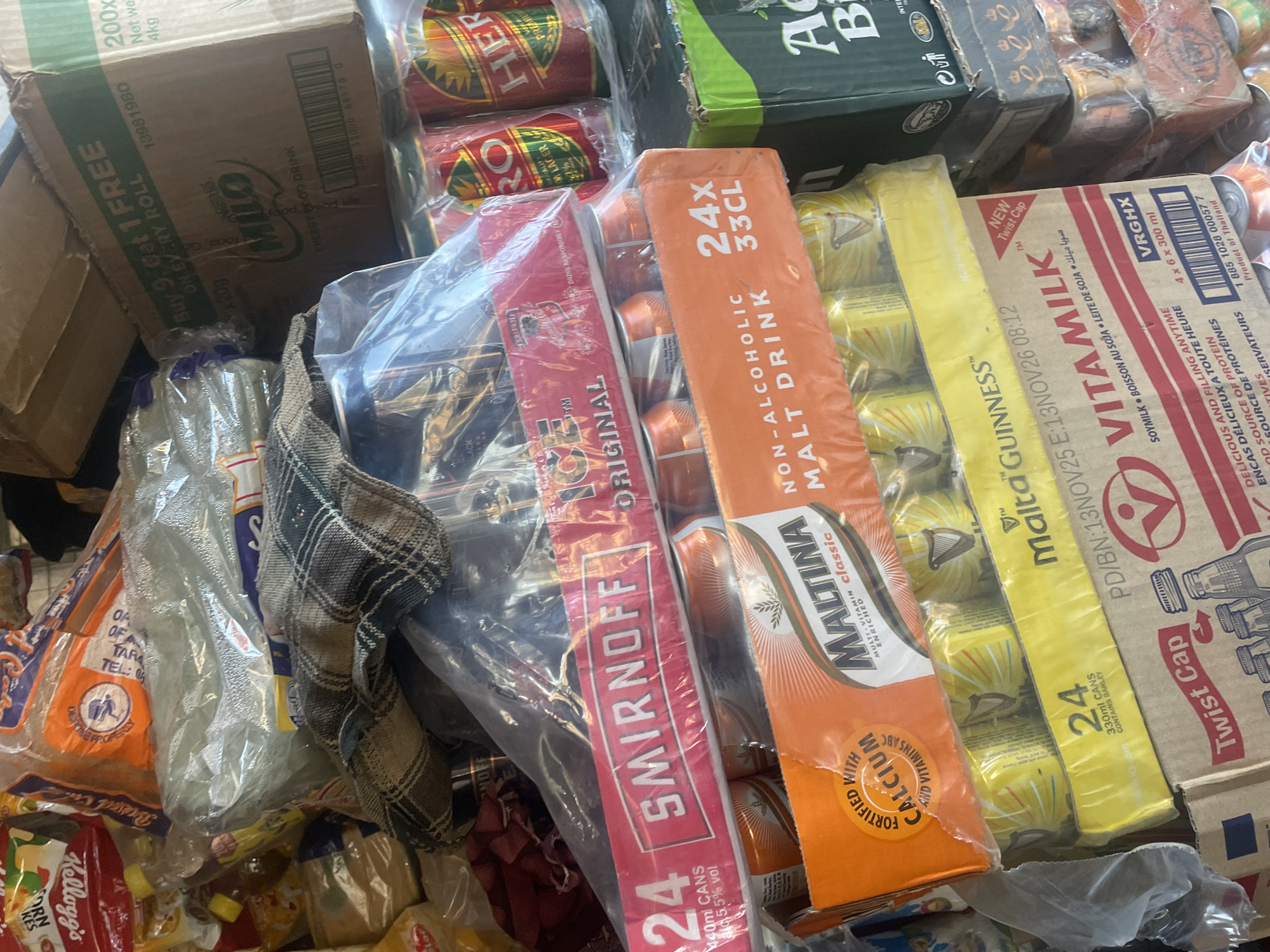 The image depicts a cluttered pile of various beverages and food items, likely in a storage area or a market stall in Jalingo, Nigeria. Prominently displayed are several multipacks of canned drinks, including Smirnoff, Maltina, and VitaMilk. There are also boxes of Nestlé Milo energy drink and what appear to be cans of Hero Lager. Some of the packaging is plastic-wrapped, and a checkered fabric is draped over some items. The background is obscured by the density of the stacked goods, but hints of cardboard boxes and plastic sheeting are visible. The overall impression is one of disarray and abundance of merchandise.
