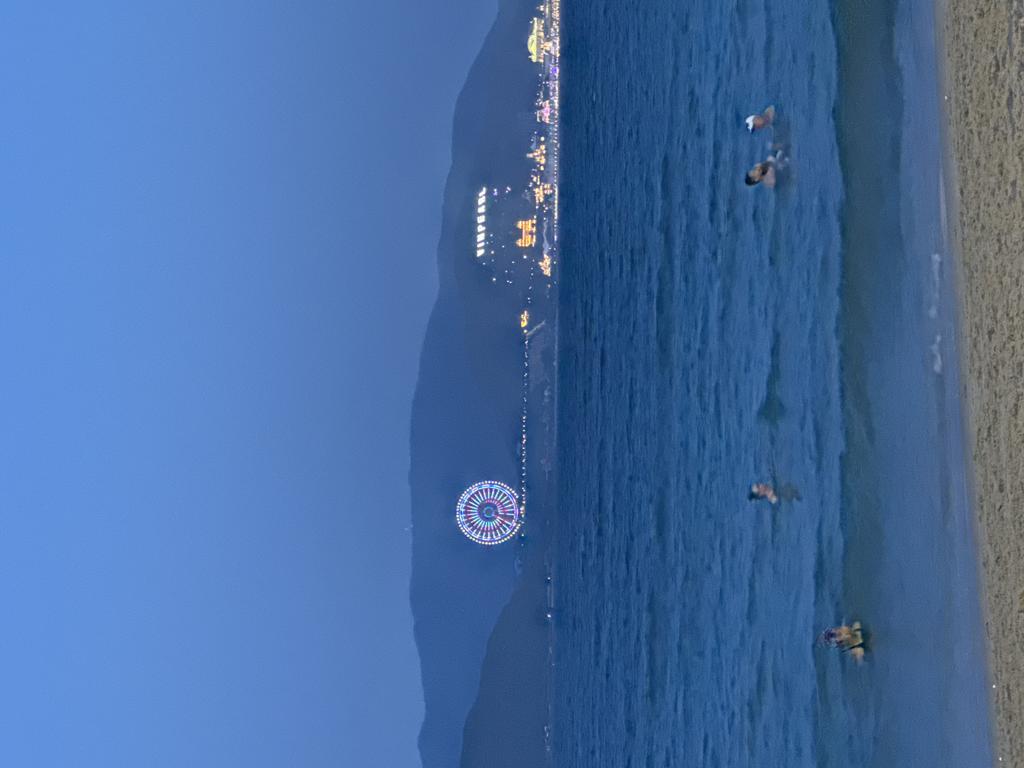 This image depicts a nighttime or dusk scene viewed from the coast of Nha Trang Ward, Vietnam, overlooking Hon Tre Island.
In the background, a range of dark mountains rises against a deep blue sky. On the left side of these mountains, a large, circular Ferris wheel is brightly illuminated with multi-colored lights in radial patterns. To the right, illuminated white letters spell "VINPEARL" across the mountainside. Below these letters and extending to the right are numerous brightly lit buildings and structures, indicative of a large resort or entertainment complex. Small lights line the base of the mountains between the Ferris wheel and the "VINPEARL" sign.
The midground consists of the dark blue ocean, characterized by gentle waves with visible white caps near the shore.
In the foreground, a strip of light brown sand, presumably a beach, is visible at the bottom of the frame. Several individuals are present in the ocean. On the far left, one person is partially submerged, facing left, with their head and shoulders above the water. Near the center-left, another individual is swimming, with their head and shoulders visible. On the right side of the frame, two individuals are together in the water, both partially submerged. One of these individuals appears to be wearing a light-colored cap.