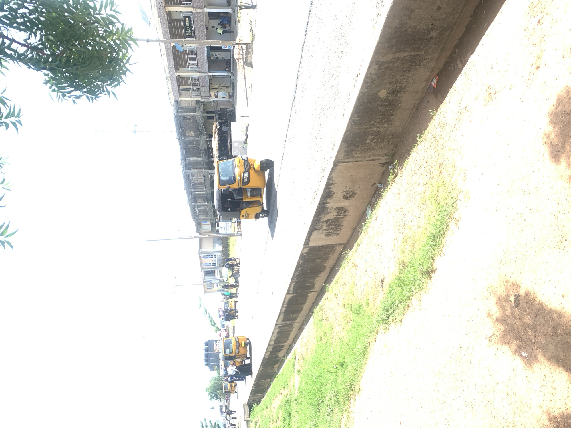 The image depicts a street scene in Jalingo, Nigeria, with a prominent focus on several yellow auto-rickshaws, commonly known as Keke Napeps. The vehicles are lined up along a paved road, suggesting a transportation hub or a busy thoroughfare. Buildings are visible in the background, indicating an urban or semi-urban setting. The lighting suggests it is daytime, likely with bright sunlight given the overexposed areas.
The primary subjects are the Keke Napeps, some of which appear to be occupied by people. The scene suggests general activity related to public transportation, with people potentially waiting for or alighting from the vehicles. The overall atmosphere appears to be one of everyday life in a Nigerian city.
Notable details include the color of the Keke Napeps, which are consistently yellow, a common color for such vehicles in the region. The road surface is concrete, and a grassy area with some sparse vegetation is visible to the right of the road. There is some debris on the ground, including a small red and white packet. A sliver of a tree with green leaves is visible in the top left corner.
The image is oriented vertically, suggesting it was taken with the camera held in portrait mode. The composition shows a line of buildings on the left and the road and accompanying embankment on the right, with several Keke Napeps parked or moving along the road. The presence of multiple vehicles and buildings indicates a populated area. There is some faint text visible on one of the Keke Napeps, but it is not legible.