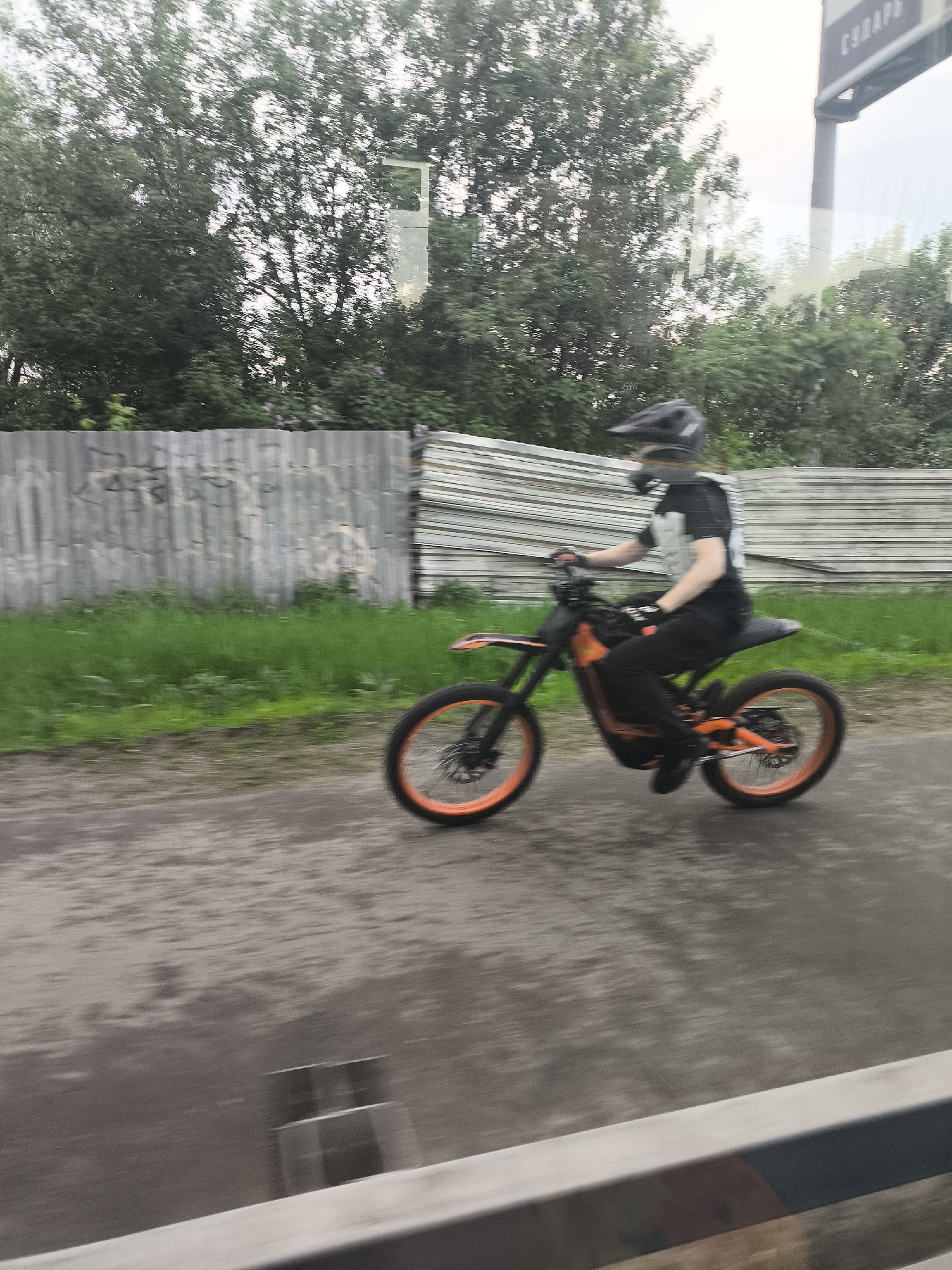 A blurry, eye-level shot depicts a person on an electric dirt bike driving on a wet, dark road. The biker wears a dark helmet, t-shirt, and pants. The dirt bike is orange and black. In the background, a gray, corrugated metal fence is partially obscured by green grass. Over the fence is a dense green tree line with a sign visible on a metal post. The image is taken from behind a metal railing, suggesting the photographer is inside a moving vehicle. The location is Unknown, Unknown.