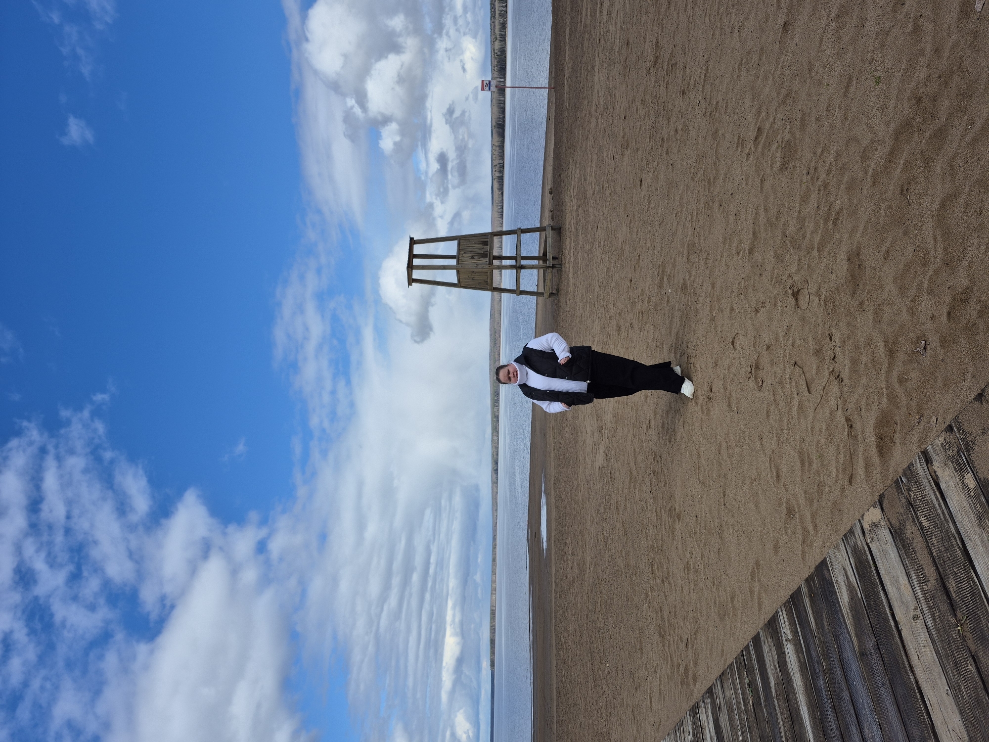 The image depicts a beach scene in Chaikovsky, Russia. The sky is partly cloudy with blue patches. A body of water stretches towards the horizon, with a sandy beach in the foreground. A wooden lifeguard tower stands on the beach. A person in dark clothing and white shoes is positioned on the beach, near the shoreline. Wooden planks are visible in the lower right corner of the image.