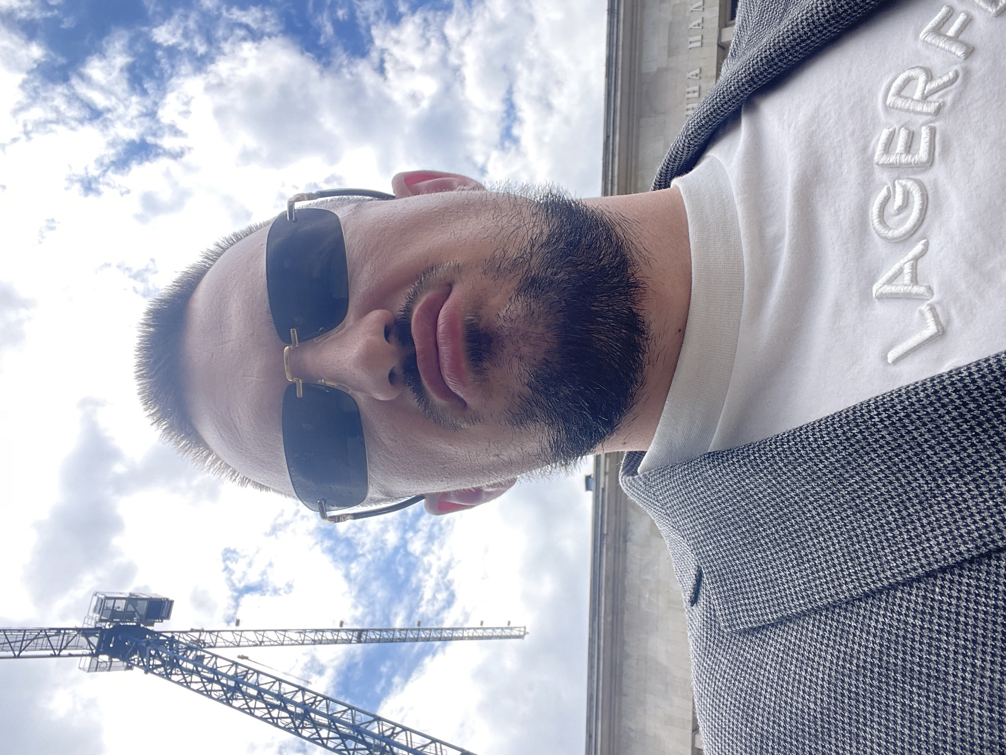 A selfie-style photograph captures a man in front of a building and a crane. The man has dark hair and a beard, and he is wearing sunglasses, a white t-shirt with "LAGERFEL" visible on the front, and a grey herringbone jacket. The building behind him appears to be a large, stone structure. To the left, a construction crane is visible against a cloudy sky. Based on the architectural style and the Cyrillic text that is partially visible on the building, the location is likely in Sofia, Bulgaria, specifically at the National Palace of Culture (NDK).