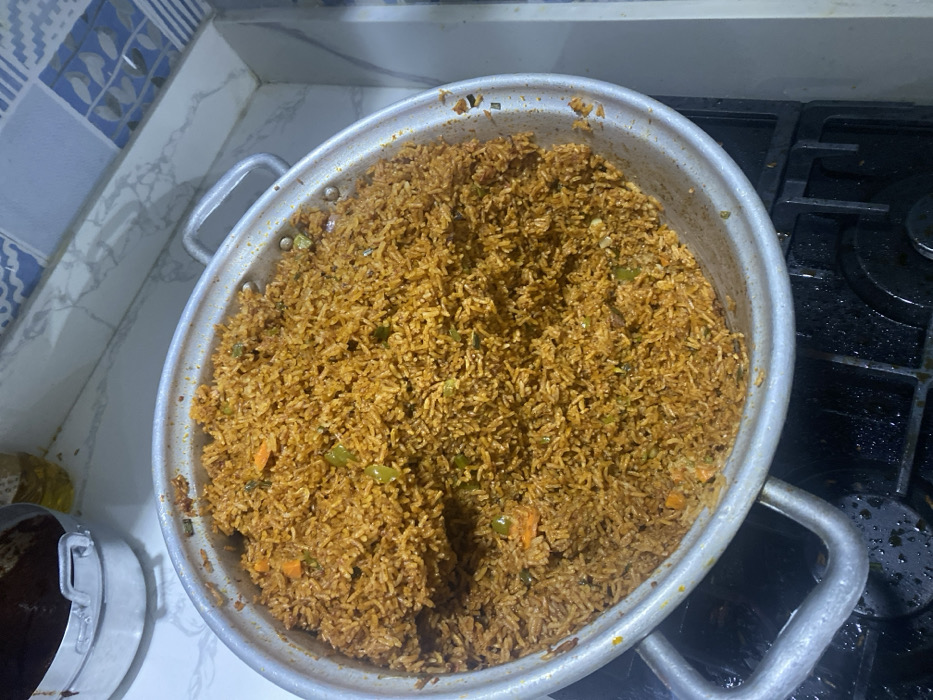 A large metal pot brimming with cooked Jollof rice is the central subject of this overhead shot, likely taken in a kitchen in Adenta Municipality, Ghana. The rice displays a characteristic reddish-brown hue, interspersed with visible pieces of chopped carrots and green bell peppers. The pot itself is silver-colored, with two sturdy handles, suggesting it's a sizable vessel for cooking large quantities.
The pot is situated on a kitchen surface, with the left side resting on a white, marble-patterned countertop. A blue and white patterned tiled backsplash is visible behind this section of the counter. The right side of the pot extends over a black gas stovetop, which has visible grates and some minor cooking residue, indicating it has been recently used. A portion of another metallic pot or container is faintly visible on the far left of the image. The scene conveys the preparation of a hearty meal, typical of home cooking. There is no visible text in the image.