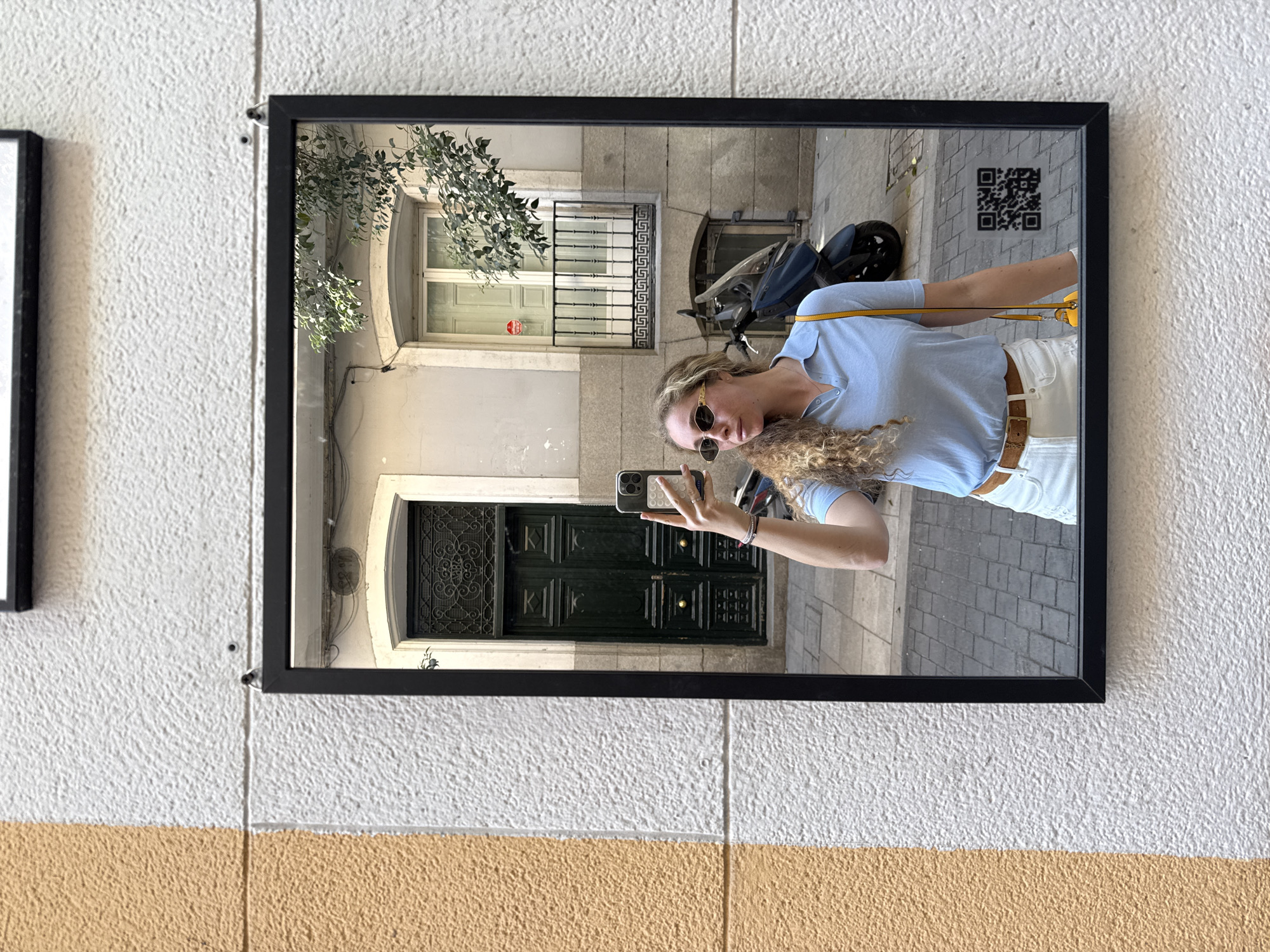 The image shows a rectangular, black-framed mirror hung on a textured white and tan wall, presumably outside. The reflection in the mirror depicts a street scene in Madrid, Spain. A woman is taking a selfie with her phone, reflected in the mirror. She is wearing sunglasses, a light blue short-sleeved shirt, white pants with a brown belt, and a yellow purse strap is visible over her shoulder.
Behind her in the reflection, a building with a dark green double door, windows with ornate metal grilles, and a parked blue scooter are visible. A QR code sticker is affixed to the building in the upper right corner of the mirror's reflection. There are some leafy tree branches visible at the top left of the reflection.
The wall the mirror is hanging on is split horizontally, with a white, stucco-like texture above a tan section. Another, smaller black-framed object is partially visible on the wall to the left of the mirror. The photo is taken at an angle, slightly skewed.