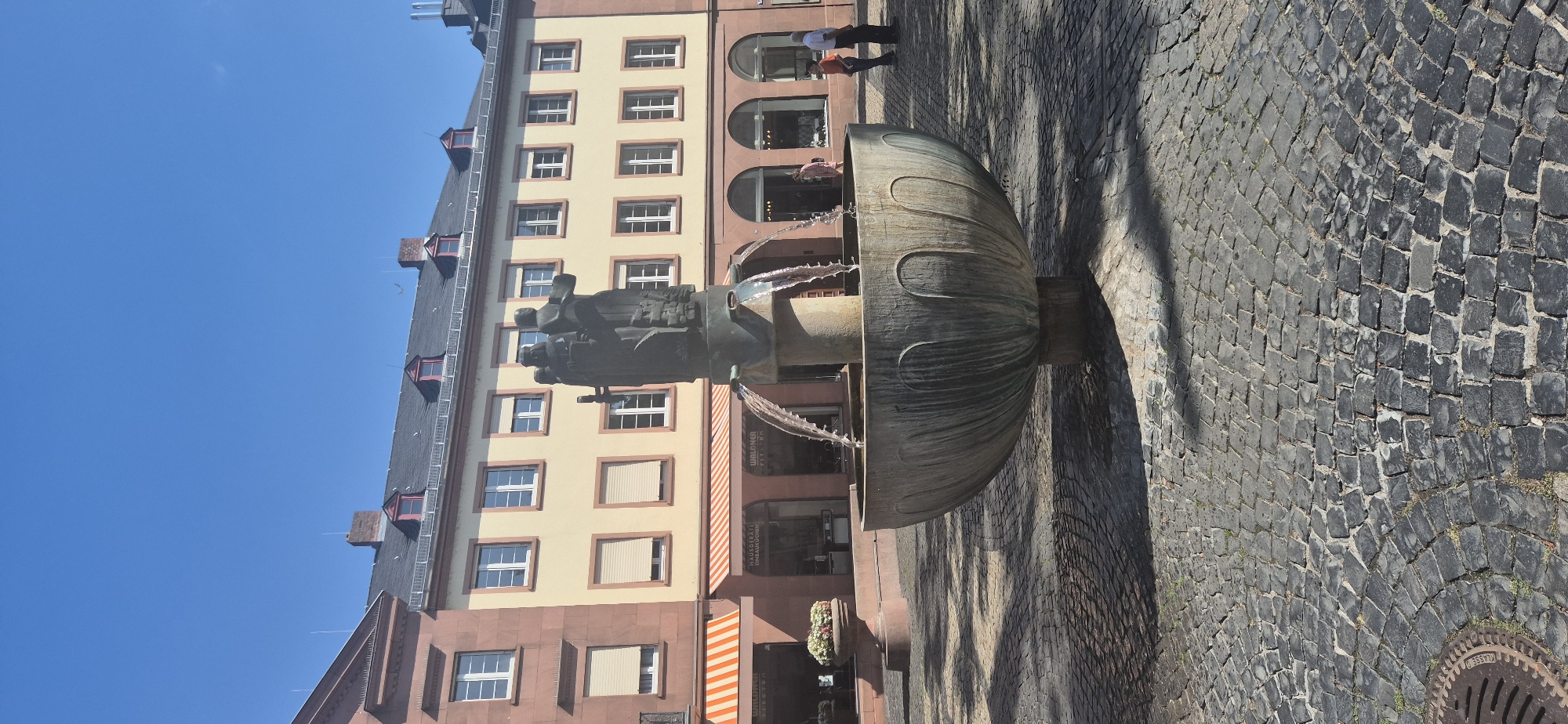 This is a full-frame, eye-level, vertical shot of a fountain in a cobblestone square in Mainz, Germany, specifically the market square in front of St. Christoph's Church. The fountain, made of dark stone with a weathered patina, features a sculptural element of figures at its apex. Water cascades from the upper part of the fountain into a large, wide bowl.
Behind the fountain, a building with a yellow facade and red-tiled roof dominates the background. This building has multiple windows with beige rolling shutters, and a distinctive red-brick section on the left. Some shops are visible at the ground level, with striped awnings.
In the middle ground, two individuals stand near the shops. A woman in a pink dress is partially visible near an arched entrance, and a man in a white shirt and dark pants stands further to the right, next to another man in an orange shirt and dark pants.
The foreground consists of a textured cobblestone ground, with visible shadows cast by the fountain, suggesting it is a sunny day. A manhole cover with a circular pattern is visible in the lower left corner. The sky is clear and bright blue.