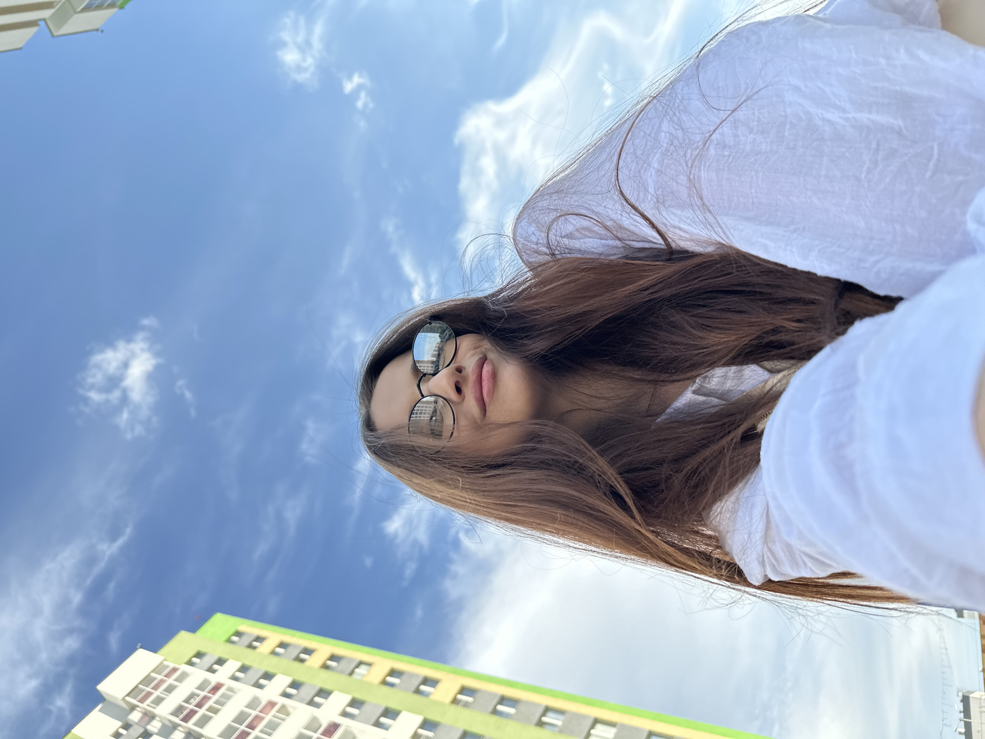 A low-angle shot captures a young woman with long brown hair and sunglasses wearing a white shirt. The background features a blue sky dotted with fluffy white clouds and the top corner of a modern, multi-story building with green and yellow accents. The woman's face is angled towards the camera, revealing her lips and nose. The style of the building suggests a location in a contemporary urban environment. The location is Yekaterinburg, Russia.