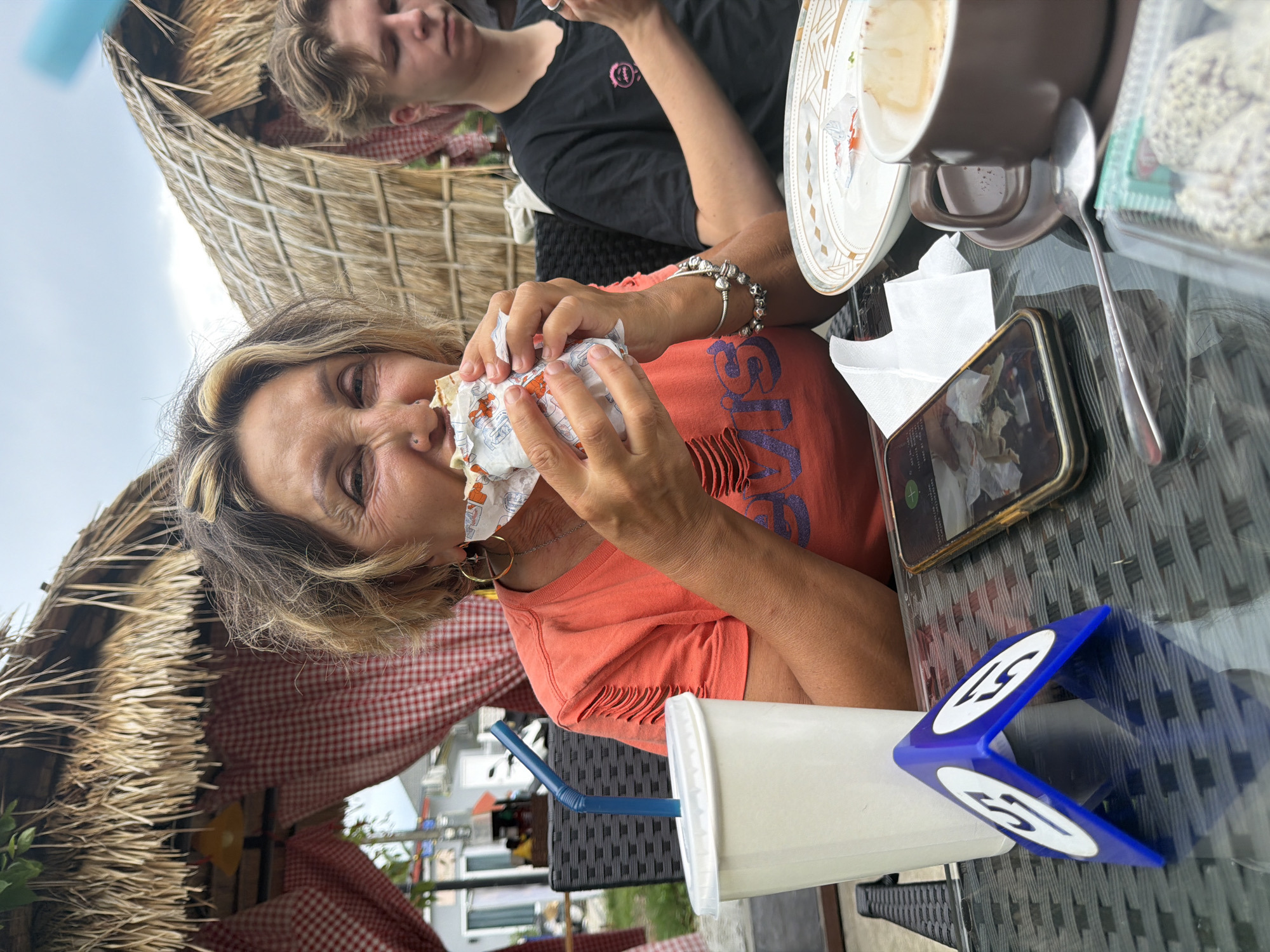 The image shows a middle-aged woman with light brown hair eating a wrapped item, likely a sandwich or burger. She wears an orange t-shirt with fringed sleeves and a silver bracelet. Her face is partially wrinkled, and she appears to be eating with gusto.
A young adult with blond hair sits across from her, partially visible in the background, wearing a black t-shirt.
A table setting includes a half-eaten plate, a coffee cup on a saucer, a spoon, a container of sweets, a cell phone, a white cup with a blue straw, and a blue table marker displaying "51".
The scene appears to be outdoors, under a thatched roof structure with red-and-white checkered fabric visible. A building and some greenery can be seen in the background.
The location is identified as Choeng Thale, Thailand.
