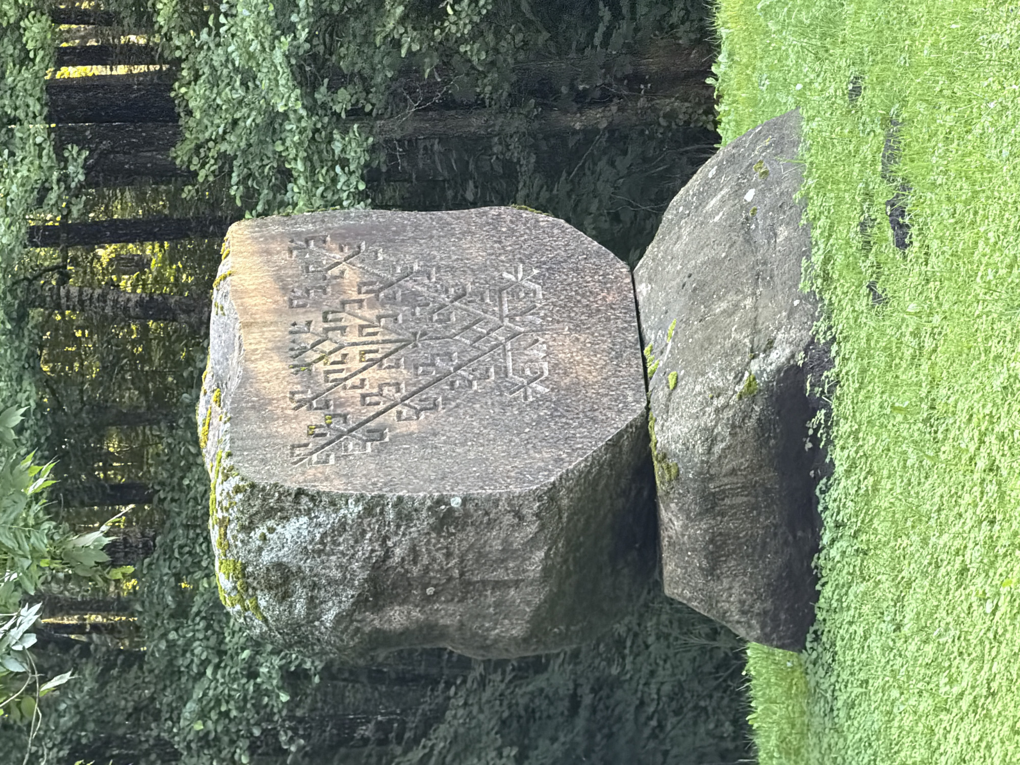 In the city of Ulbroka, Latvia, a large, ancient boulder stands as a monument. The boulder is carved with intricate patterns, resembling a tree of life or a snowflake. The carving is detailed and appears to be ancient. The boulder is situated in a grassy clearing, surrounded by lush green trees and foliage. The scene is serene and natural, evoking a sense of peace and tranquility. The boulder itself is majestic, a testament to the enduring power of nature and human artistry. It is a place where one can connect with the past and appreciate the beauty of the natural world.