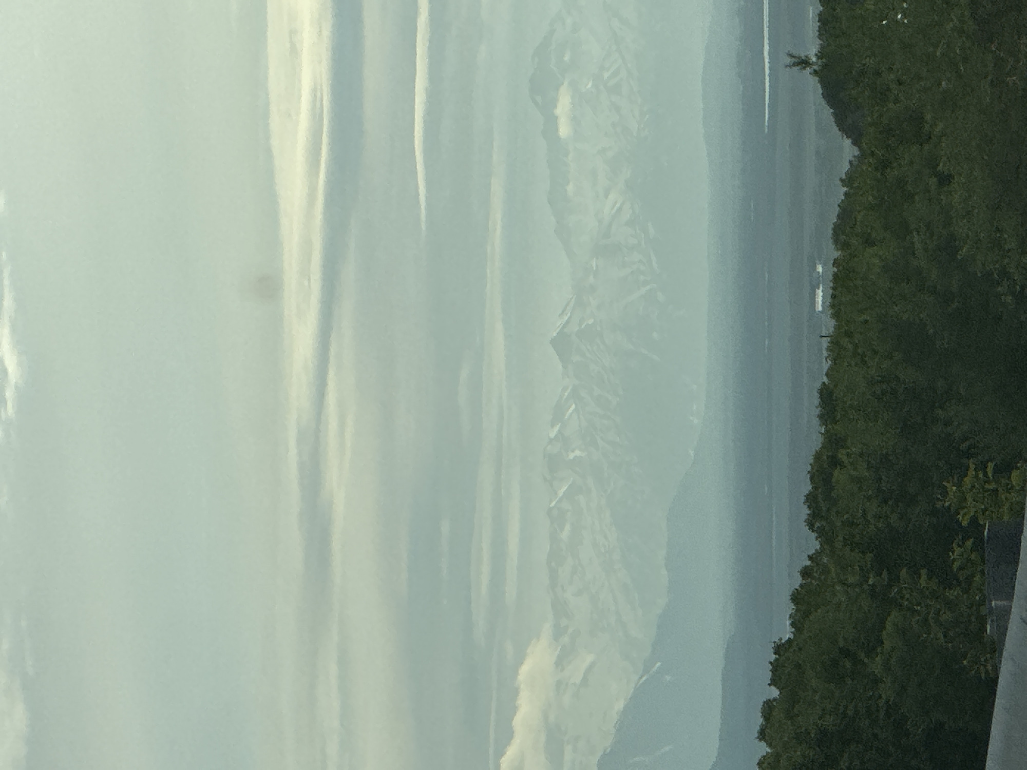 A hazy, panoramic view captures a vast landscape from an elevated position. In the foreground, a dense line of dark green trees stretches across the bottom of the frame, with the top edge of a concrete barrier visible at the very bottom. Beyond the trees, a wide, flat expanse of land is shrouded in mist, with faint outlines of buildings and a body of water visible in the distance. Towering over this plain are majestic, snow-capped mountains that recede into the hazy sky. The sky itself is a muted blue-grey, filled with soft, horizontal clouds that create layers of light and shadow across the scene. The overall impression is one of immense scale and natural beauty, with the atmospheric conditions lending a serene, almost ethereal quality to the image.