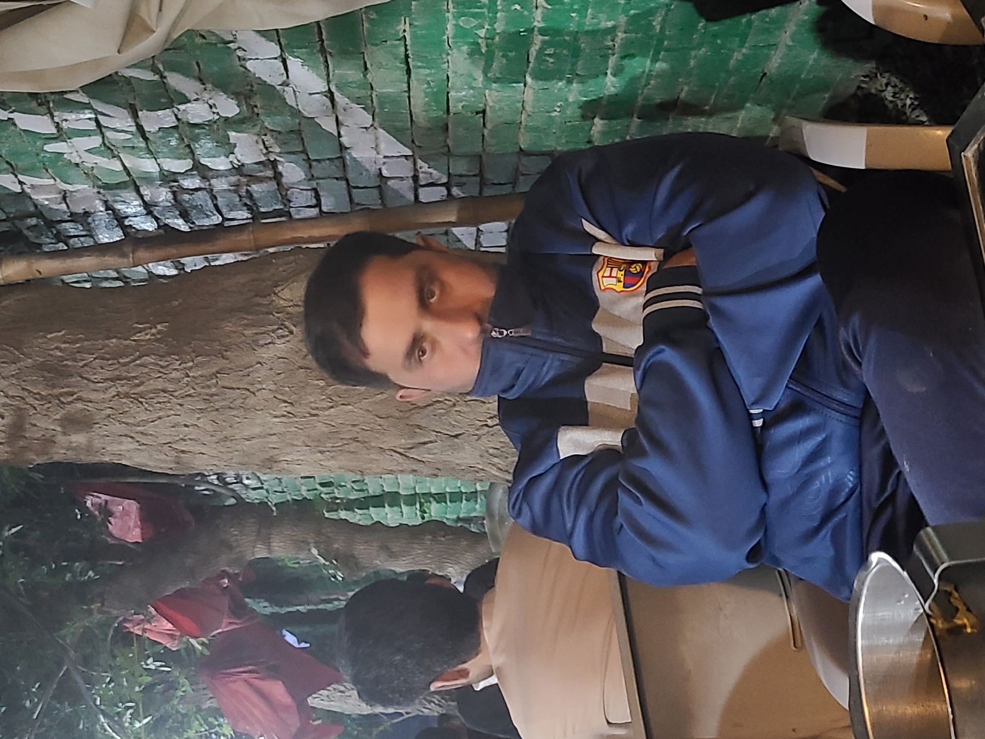 A person is sitting at a table in an outdoor setting. The person is wearing a blue jacket and dark pants. The setting appears to be in Quetta City Tehsil, Pakistan, based on context. The location is possibly a restaurant or cafe with an overhead structure of green-painted bricks and bamboo. Other people are present in the background, and there are trees and foliage in the periphery.