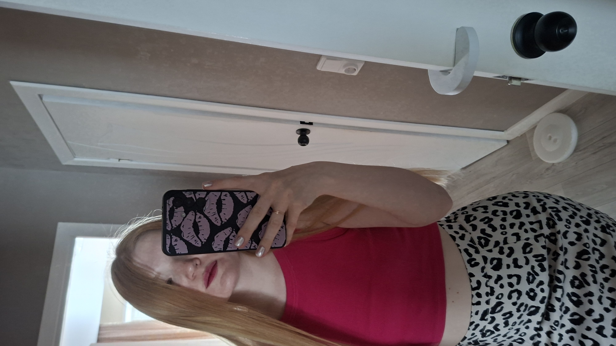 A woman is taking a selfie in a room. She is wearing a bright pink top and a white and black leopard print skirt or pants. She is holding a phone with a black case covered in purple lip prints. Her hair is long and blonde.
The room appears to be a bathroom. A white door with a black knob is visible. Above the door is a beige area with a motion sensor. Above that is a horizontal surface, likely a shelf. There is a white hook, a black doorknob, and a white decorative object resembling a button on the shelf. The walls appear to be painted a light color. The floor is wood-patterned.
The photo was taken inside, the location is unknown.