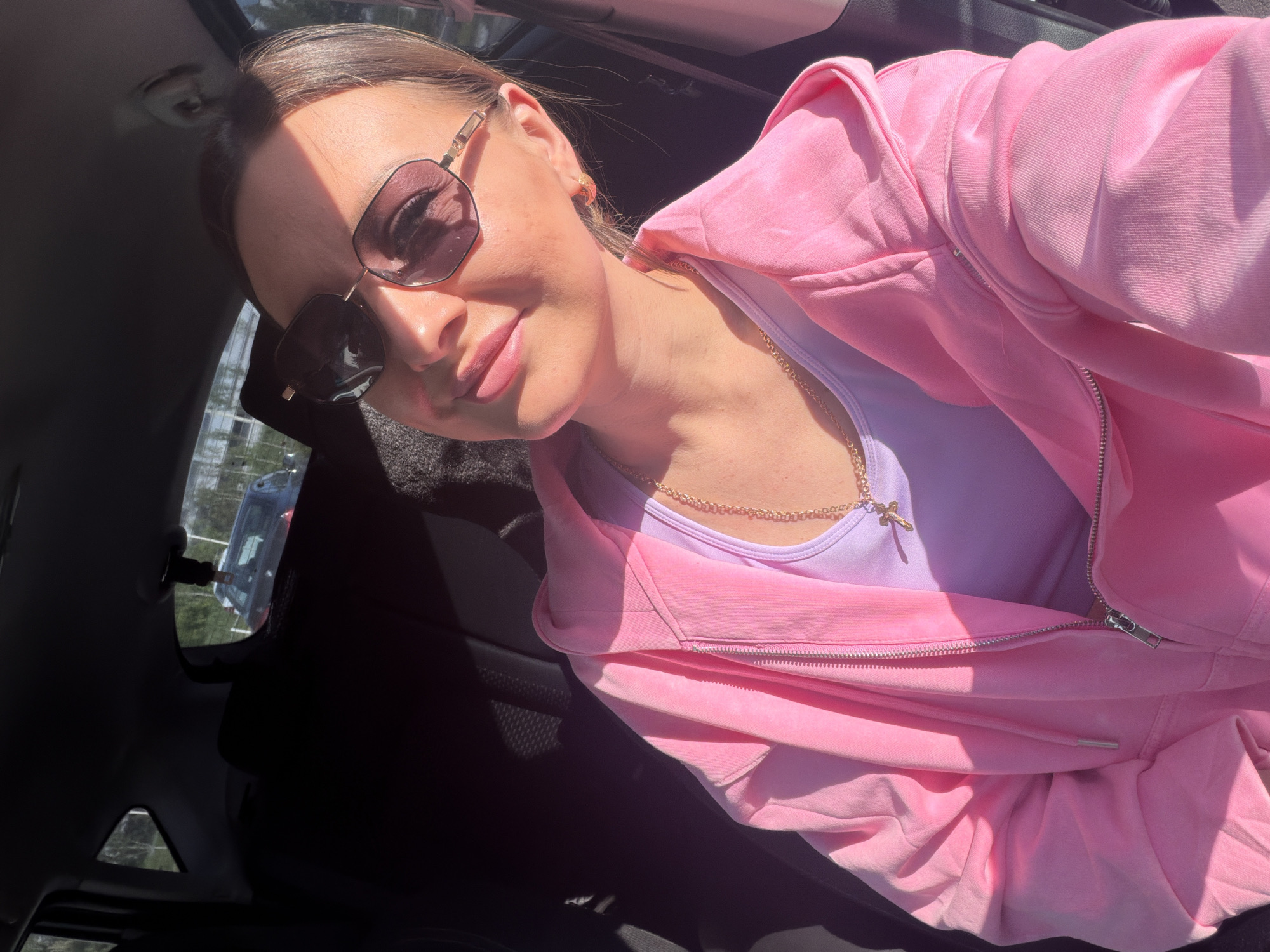 A woman is taking a selfie inside a car. She is wearing sunglasses, a lilac tank top, and a pink zip-up hoodie. A gold chain with a cross pendant hangs around her neck. Light streams in through the car windows. The location is not discernible from the provided image.