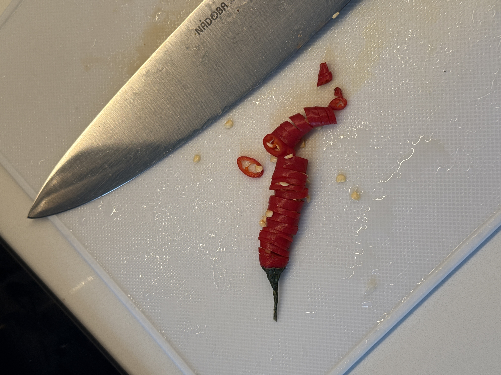 An overhead close-up shot depicts a culinary preparation scene in what appears to be a kitchen. The primary subjects are a large chef's knife and a sliced red chili pepper, both resting on a white plastic cutting board.
The metallic chef's knife is positioned diagonally across the top left quadrant of the frame, with its blade angled towards the lower center. The brand name "NADOBA" is clearly visible, etched into the upper part of the blade. Below "NADOBA," partially obscured text that seems to read "chef" or similar is also present. The blade shows signs of use with slight scratches and reflections from ambient light.
To the right of the knife, a red chili pepper has been uniformly sliced into numerous thin, bright red rings. These slices are arranged in a stack, largely preserving the original form of the pepper, with its dark green stem still intact at the bottom. Several pale yellow chili seeds are scattered both among the pepper slices and across the textured surface of the cutting board. A few smaller, irregularly shaped chili pieces are also visible.
The white cutting board features a distinctive textured, grid-like pattern across its surface. Small droplets of liquid or moisture are present on the board, particularly around the sliced pepper, suggesting recent activity. A dark edge, likely a countertop or kitchen surface, is visible along the bottom and left side of the cutting board. The scene captures a functional moment of food preparation in Shirkovo, Russia.