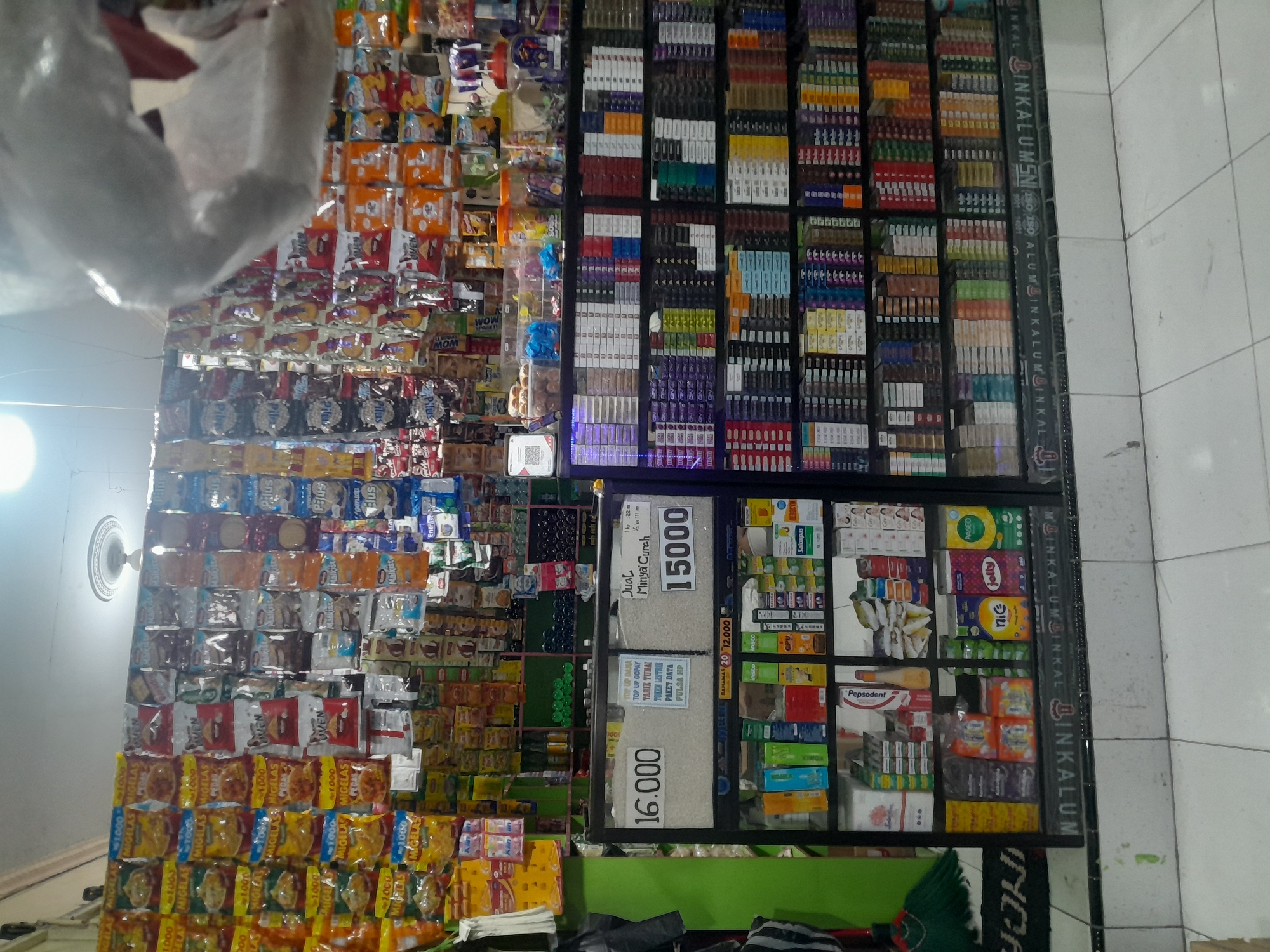 The image shows a small convenience store in Pengabean, Indonesia, stocked with various goods. The store features shelves and hanging displays filled with snacks, drinks, cigarettes, and toiletries. A glass-fronted cabinet in the foreground displays items like tissue boxes, toiletries, and small packaged goods. Prices for rice and cooking oil are displayed on signs. The store appears well-organized and densely packed with merchandise, suggesting a typical local shop catering to everyday needs. The lighting is artificial, typical of an indoor commercial space.