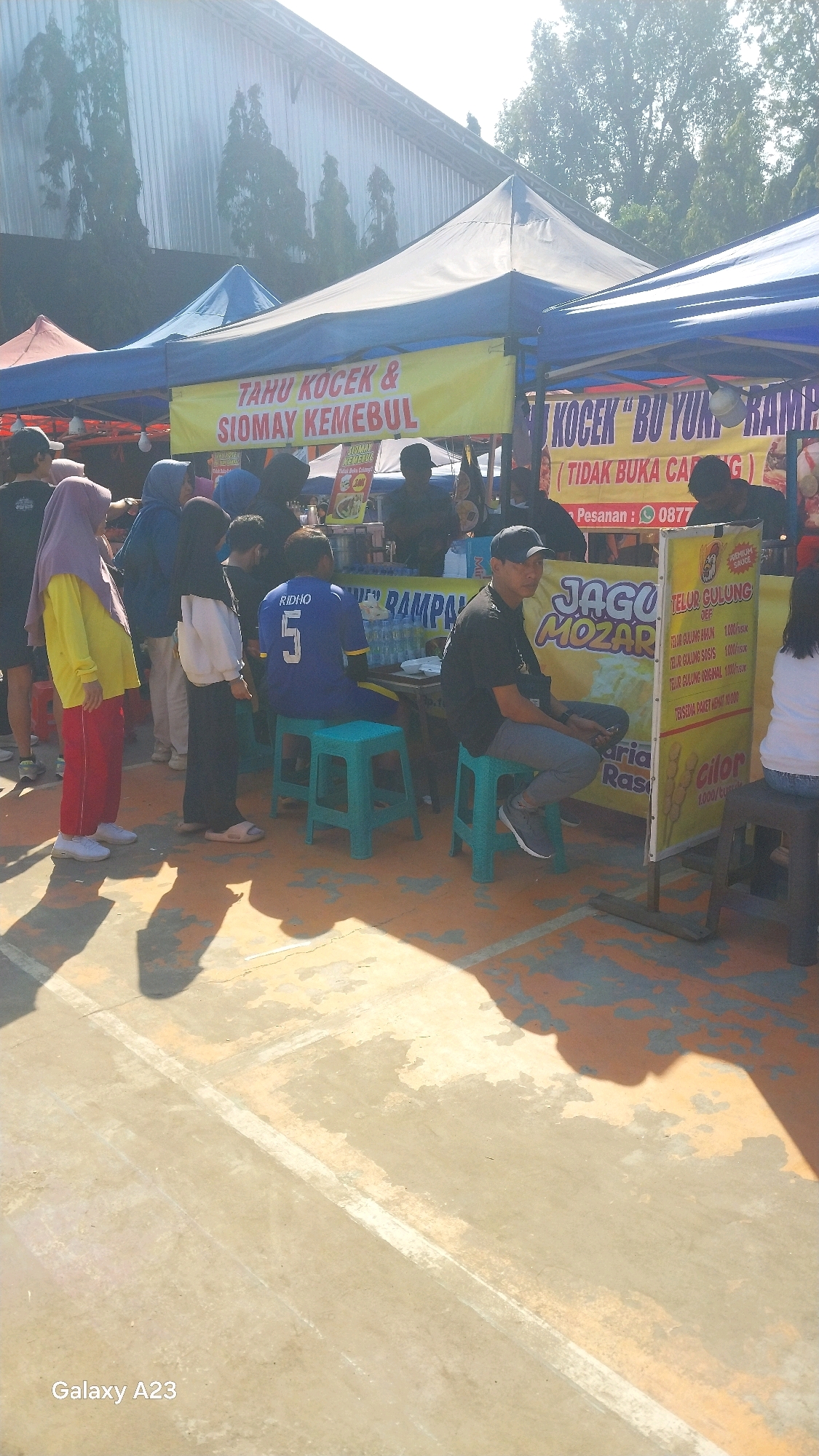 In Malang, Indonesia, a sunny outdoor scene features several market stalls. In the foreground, a concrete ground with painted lines and shadows suggests a public space. To the left, a person wearing a yellow hijab and red pants stands near a stall. Further into the image, people are seen interacting with vendors at different stalls. One stall has a yellow banner with Indonesian text that appears to advertise food items such as "TAHU KOCEK & SIOMAY KEMEBUL". Another stall prominently displays "JAGU MOZAR" and information about "TELUR GULUNG" (rolled eggs). The background reveals a large building with a corrugated metal roof and trees. The overall atmosphere suggests a lively market or food event under bright daylight.