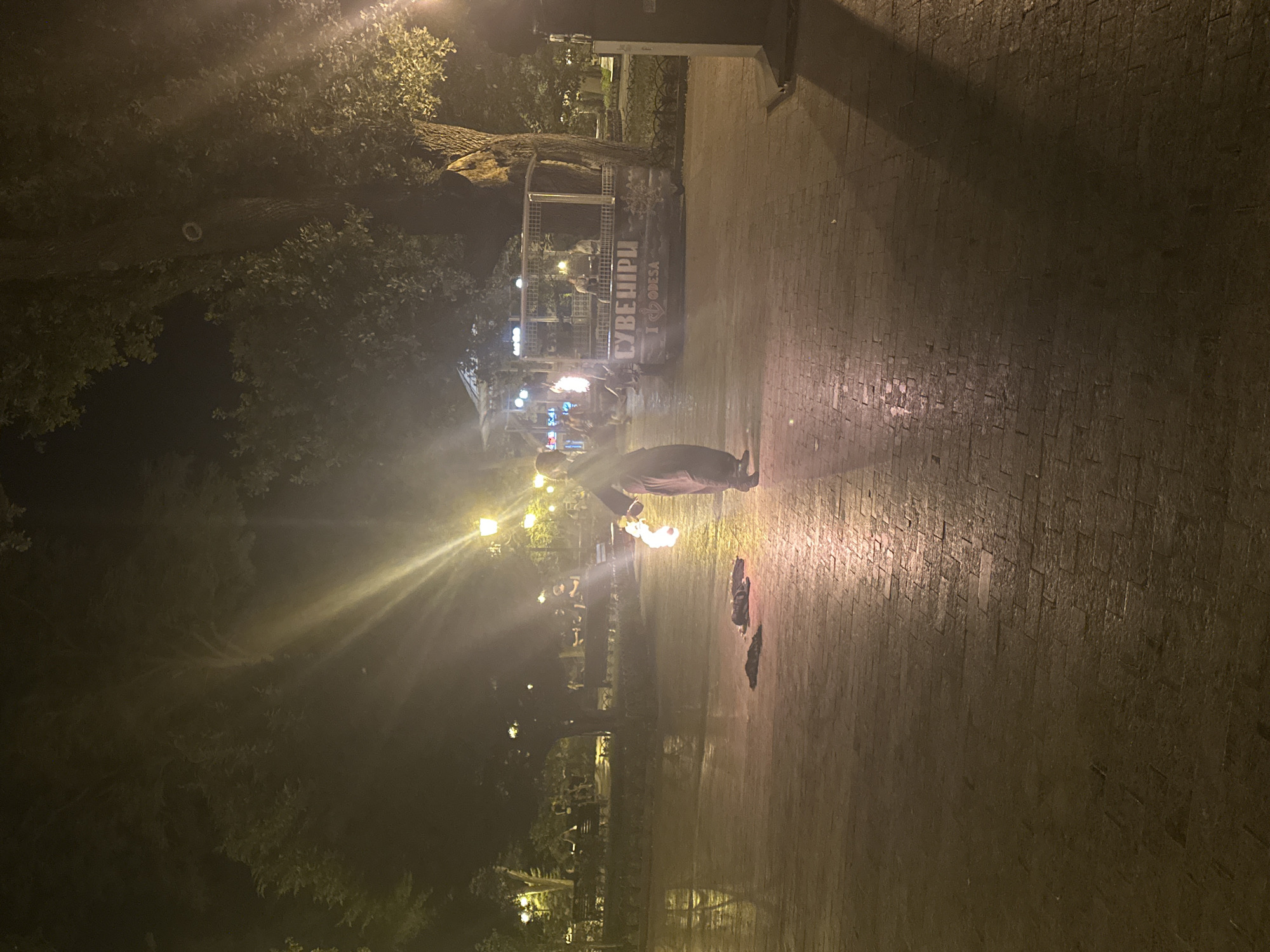The image depicts a nighttime scene in Odesa, Ukraine, likely in a park or public square. A person in dark clothing is performing with fire, holding a flaming object in their right hand. The ground is paved with bricks and appears wet, reflecting the light from street lamps. In the background, there are trees, benches, and people seated, suggesting a public gathering area. A structure with signage that reads "СУВЕНІРИ" and "I LOVE ODESA" is visible to the right, indicating a souvenir stand or shop. The overall ambiance is one of outdoor entertainment or leisure.