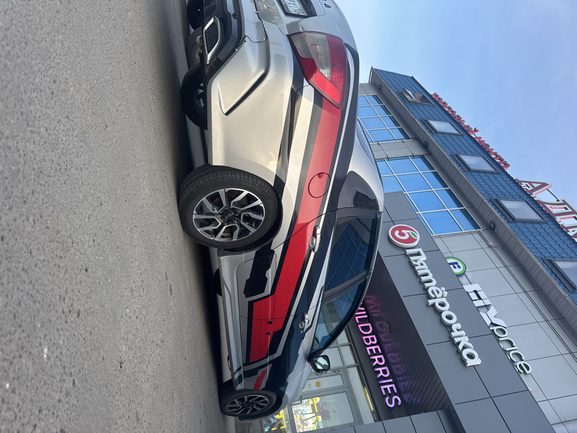 A silver car with a red and black racing stripe design is parked on a gray paved surface in front of a building with signs for "Pyaterochka" and "Wildberries." The building has blue windows and a blue roof with a red sign on top. The car has a license plate that ends in "48." The sky is blue. The location is in Chaikovsky, Russia.