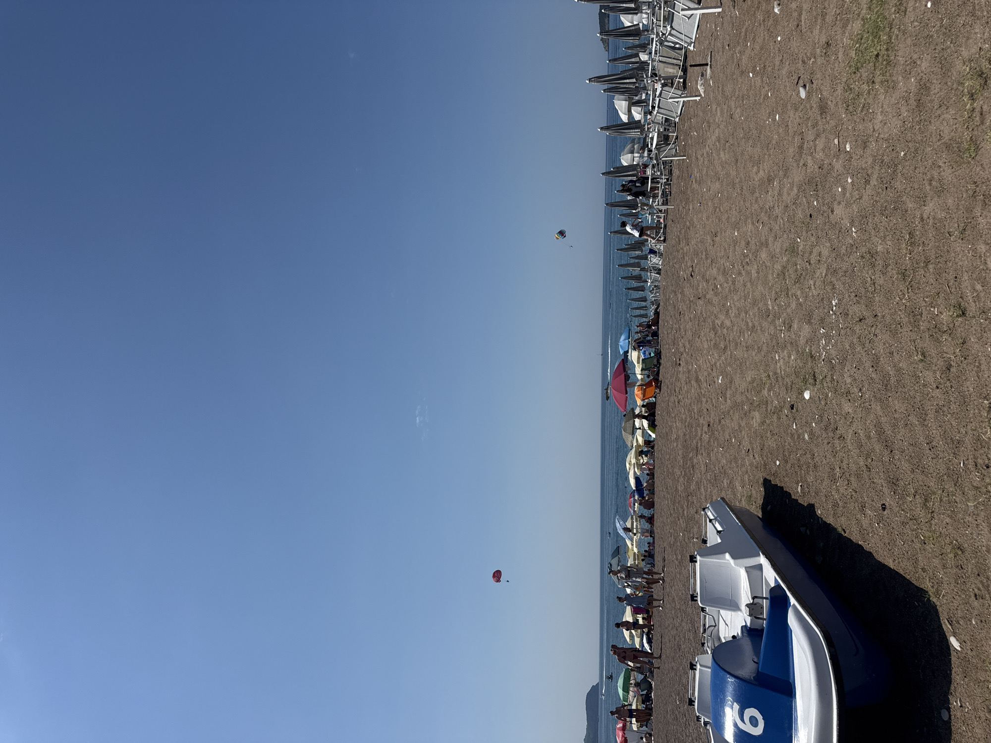 This media file captures a sunny day at a beach in Boreti, Montenegro. The scene is a bustling beachfront with numerous beach chairs and umbrellas set up on the dark sand. In the foreground, a white and blue boat with the number "9" visible on its side is parked on the shore, casting a long shadow.
The beach is populated with people, many of whom are walking or relaxing under the umbrellas. The ocean is visible in the background, with a clear blue sky overhead. The overall atmosphere appears to be one of leisure and enjoyment, typical of a summer vacation day.
Further out at sea, there are two paragliders visible in the sky, adding a dynamic element to the scene. One is a colorful parachute, and the other is a smaller red parachute. The water is calm, suggesting pleasant weather. The image depicts a vibrant beach setting, likely in the afternoon given the sunlight and shadows.