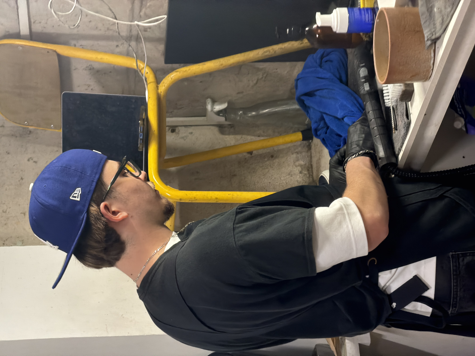 A person wearing a blue baseball cap, glasses, a black t-shirt over a white long-sleeved shirt, a silver chain necklace, and black gloves is operating a black wand-like device. The person is standing indoors, possibly in a basement or garage, near a yellow metal structure resembling scaffolding. The device is positioned near a shelf containing items such as bottles, a blue cloth, a cardboard tube, and cleaning brushes. The location is not definitively identifiable as Paris, France, based on the visual information provided.