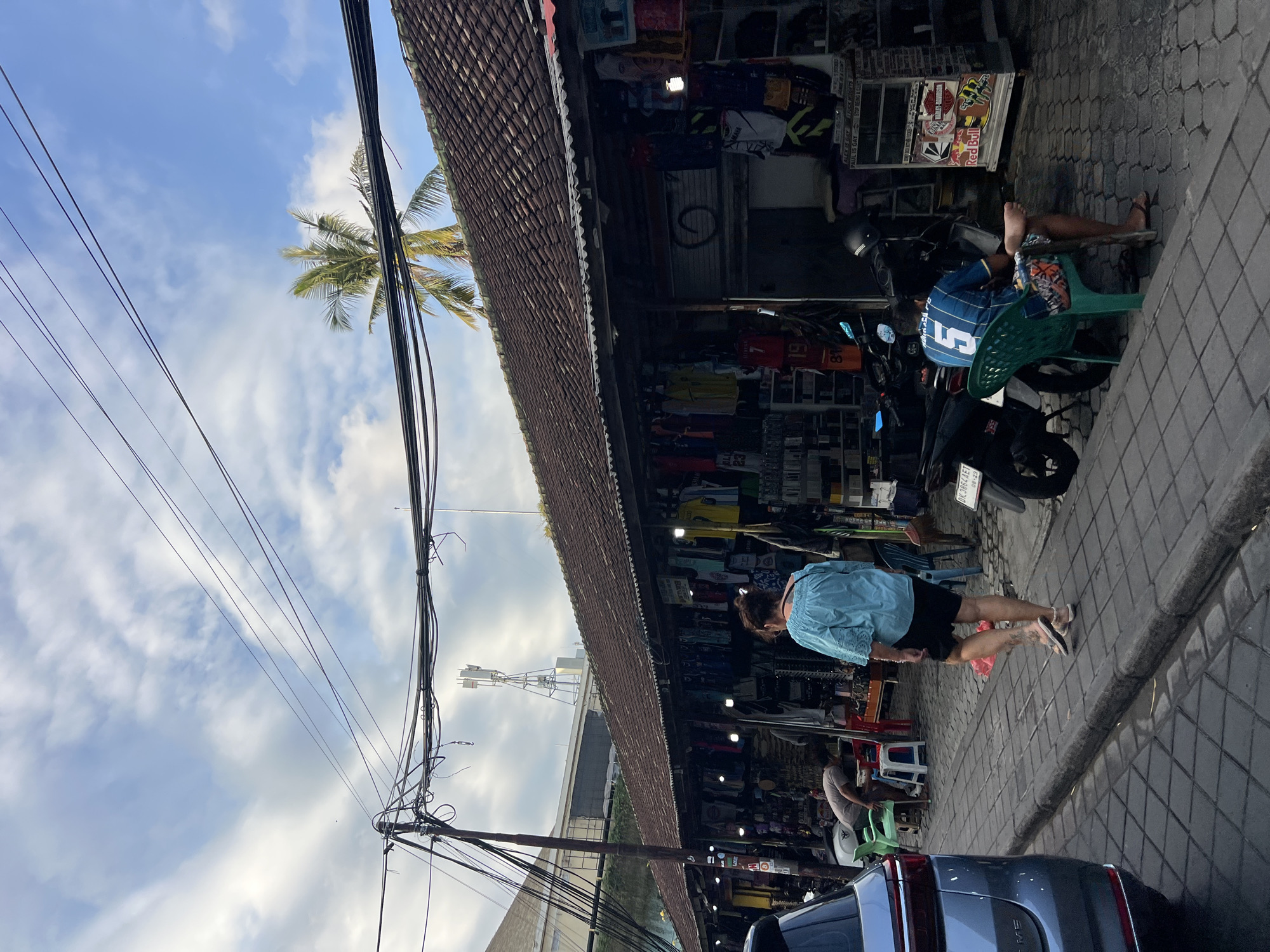In Kuta, Indonesia, a street-level view captures an outdoor market stall area with a paved walkway. A woman in a blue top and black shorts, her back to the camera, walks along the walkway. In front of her, a person is bent over near some chairs and tables. To her right, a man in a blue jersey with the number "5" sits on a green plastic chair. Several motorcycles are parked near the market stalls, which are filled with clothing and other goods. Overhead, a dense network of electrical wires spans across the sky, with a cell tower visible in the distance. A palm tree's fronds peek over the tiled roof of the market stalls. A light blue SUV is parked on the left side of the frame. The ground is paved with square tiles.