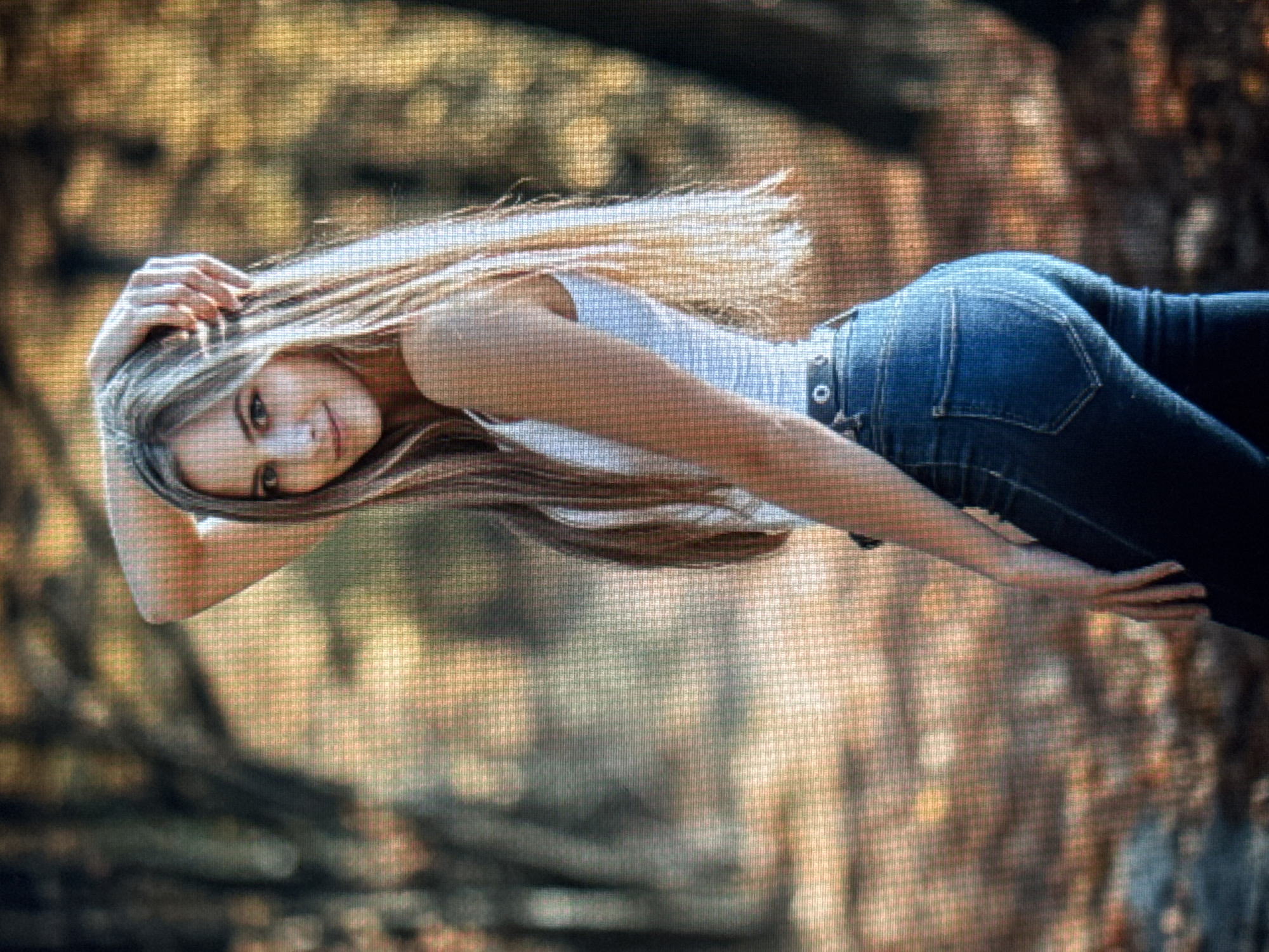 An image captures a young woman posing outdoors, horizontally across the frame. She lies on what appears to be the ground amidst blurred foliage, suggesting a natural setting like a forest or park. The woman has long, flowing light brown hair and is looking at the viewer with a warm, gentle smile, her head tilted slightly. One hand is placed near her head, resting on her hair, while her other arm extends across the frame. She is dressed in a light-colored tank top or t-shirt and form-fitting blue denim jeans with a dark belt. The background is out of focus, displaying soft, warm hues of brown, green, and gold from the surrounding trees and natural elements, hinting at an autumn or late summer atmosphere. Natural, soft lighting illuminates the scene. No specific text is visible within the image. While the provided context indicates Varna, Bulgaria, the visual elements themselves do not offer distinct geographical markers beyond a generic outdoor environment.