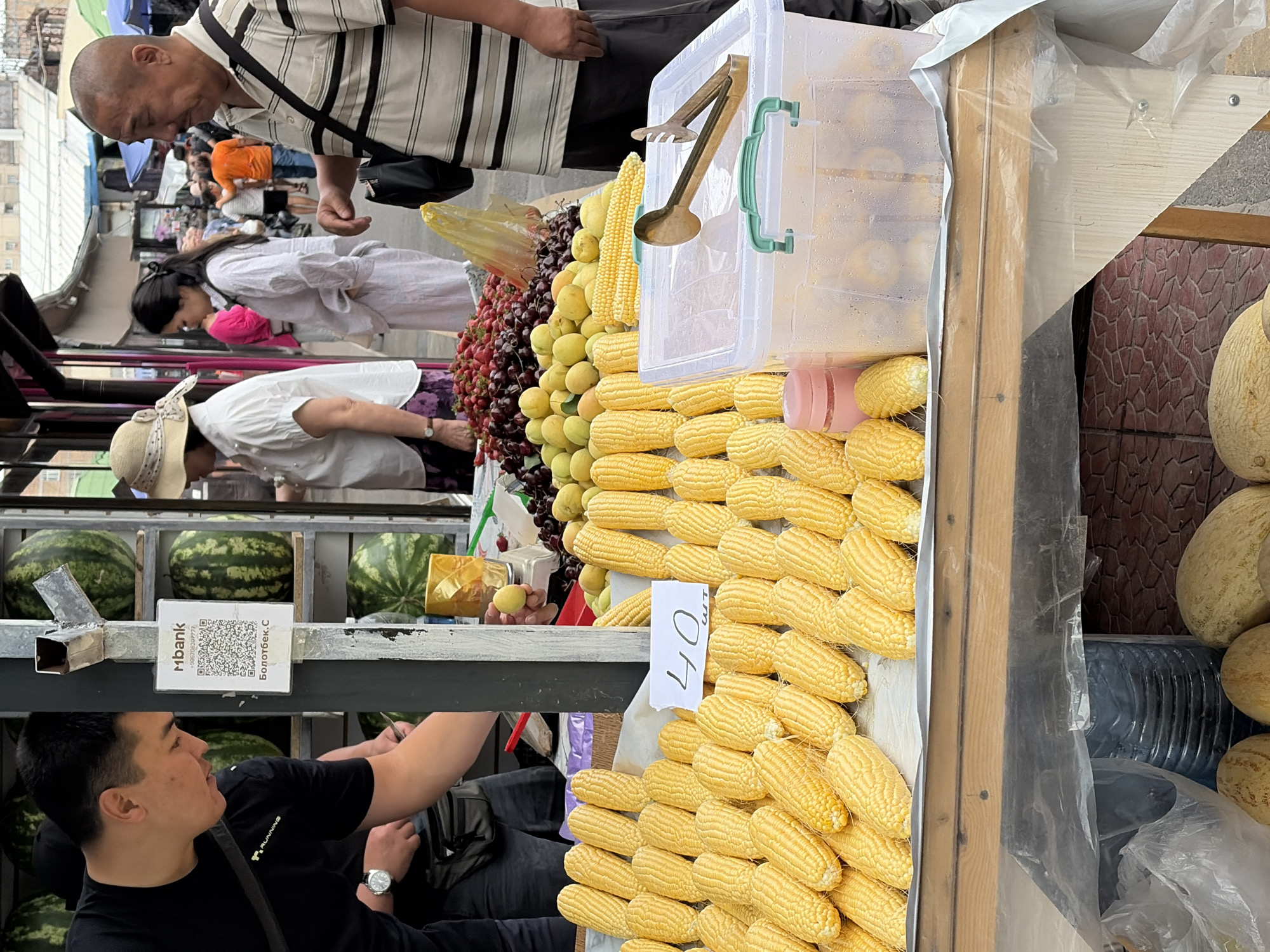 In an outdoor market stall, a man in a black t-shirt and a man in a striped polo shirt stand near a display of fresh produce. The man in black is bent over, his left arm out of frame, while the man in the striped polo shirt looks to his right. In front of them, rows of yellow corn cobs are arranged on a wooden table covered with white paper. A sign with "40" and Cyrillic characters is placed among the corn. To the right, a clear plastic container holds more corn, and a pair of metallic tongs rests on its lid. Behind the corn, a variety of fruits including strawberries, cherries, apricots, and peaches are piled high. In the background, several people are browsing the stalls. Further back, large watermelons are stacked. The market appears to be bustling with activity.