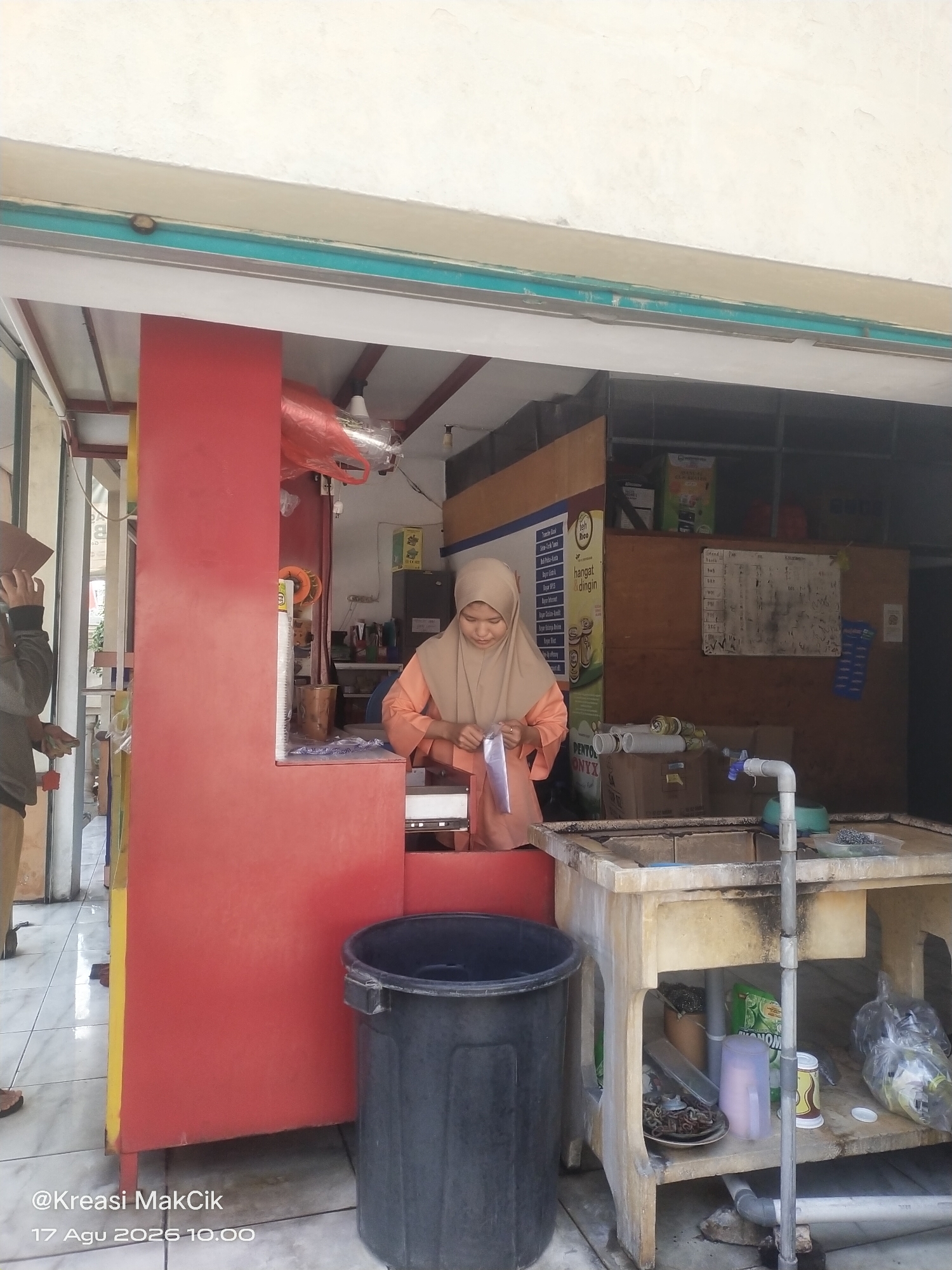 A young Indonesian woman wearing an orange hijab and an orange tunic is standing behind a red counter at an outdoor food stall in Sambi Bulu, Indonesia. She is holding a clear plastic bag and appears to be in the process of packaging an item. To her left, a large red structure, likely part of the stall's facade, is visible. In the foreground, to the right of the counter, is a concrete sink with plumbing fixtures and a grey trash bin. Various items, including boxes, bottles, and plates of food, are scattered around the sink area and on shelves beneath it. The ground is tiled. A watermark at the bottom left corner reads "@Kreasi MakCik 17 Agu 2026 10.00".