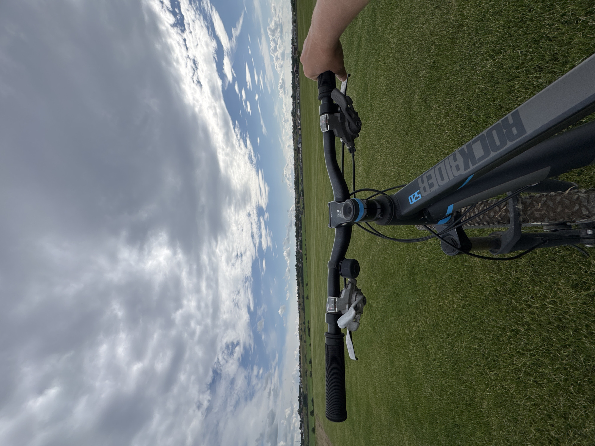 The image shows a first-person perspective from someone riding a bicycle. The bicycle's handlebars and part of the frame are visible in the foreground, with a hand gripping the right handlebar. The scene ahead depicts a wide, flat expanse of bright green grass under a cloudy sky. In the distance, a line of houses and trees forms the horizon. The location is identified as Kempelevo, Russia. The overall impression is of a scenic, open outdoor environment.