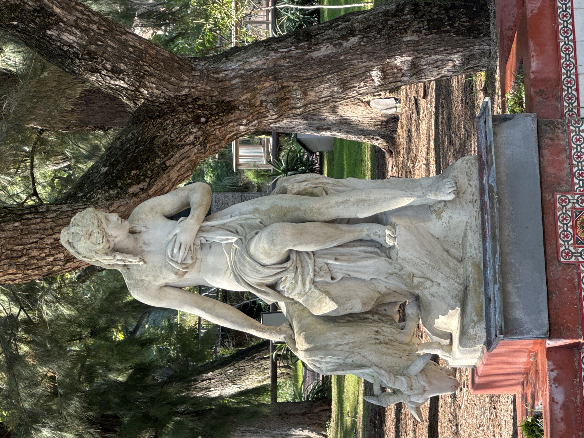 The image shows a sculpture of Europa and the Bull, located in the city of Buenos Aires, Argentina. The sculpture depicts a woman draped in cloth reclining on a bull. The sculpture is made of white marble. The base of the statue is concrete and red-tiled. There are trees behind and in front of the statue. The ground is covered in brown bark mulch.